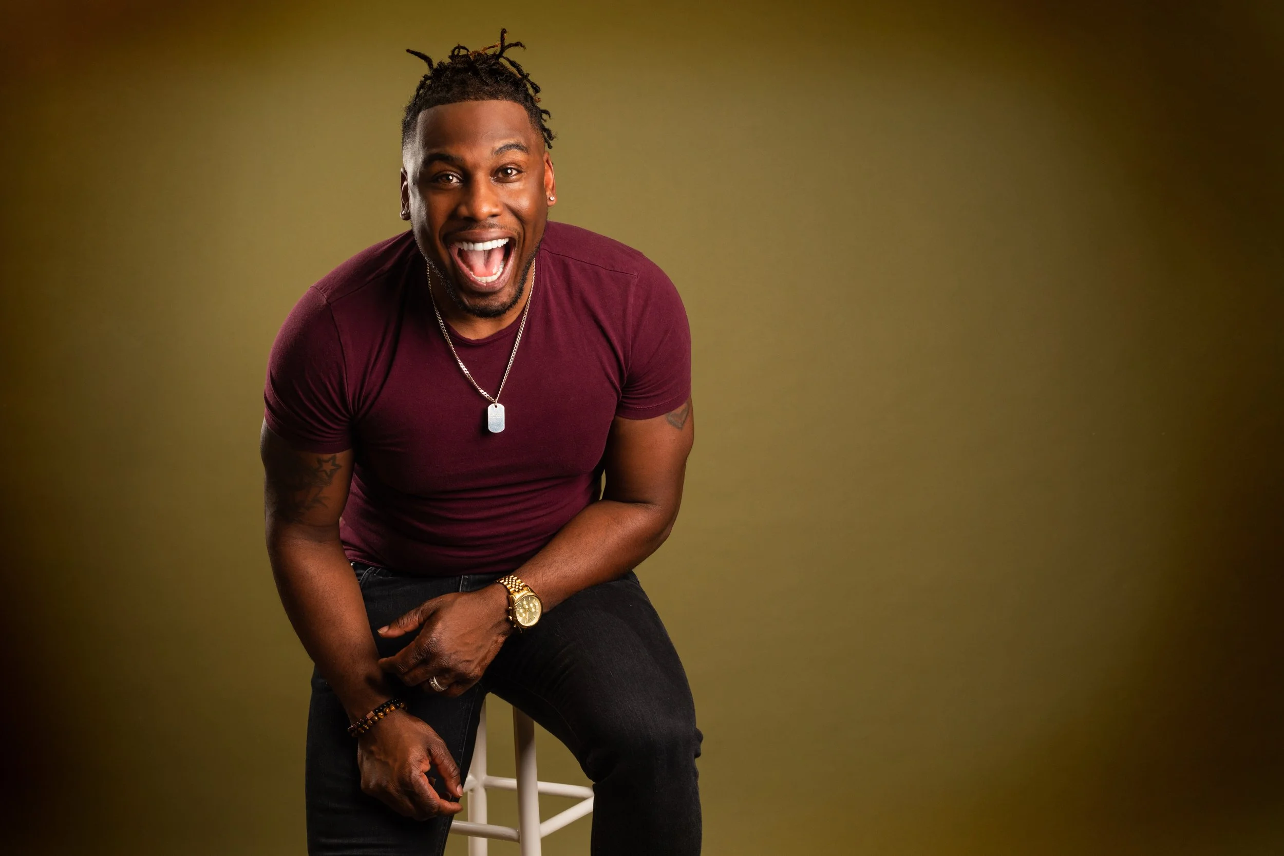 A joyful man with dreadlocks smiling and laughing while sitting on a white stool against a green background, wearing a maroon t-shirt, dark jeans, gold watch, chain, and bracelet.