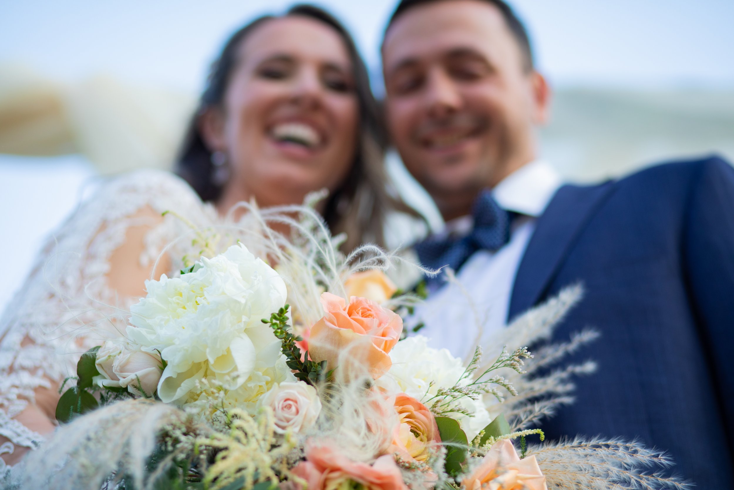 A smiling bride and groom holding a bouquet of white, peach, and pink flowers, dressed in wedding attire, with a blurred outdoor background.
