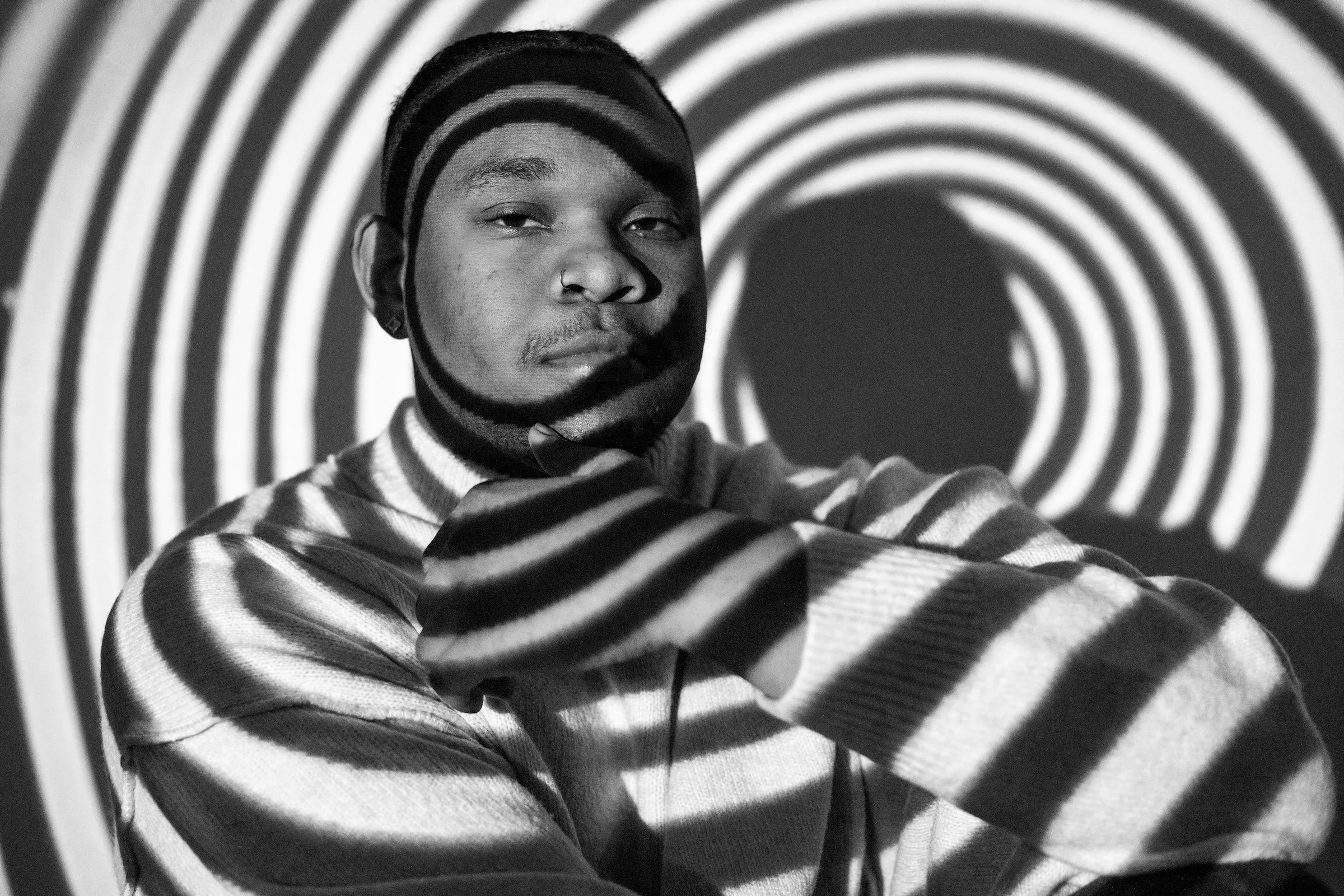 A person wearing a striped sweater and beanie hat, with the person's face and arm cast in shadow and light, against a background of concentric circles creating a hypnotic pattern.