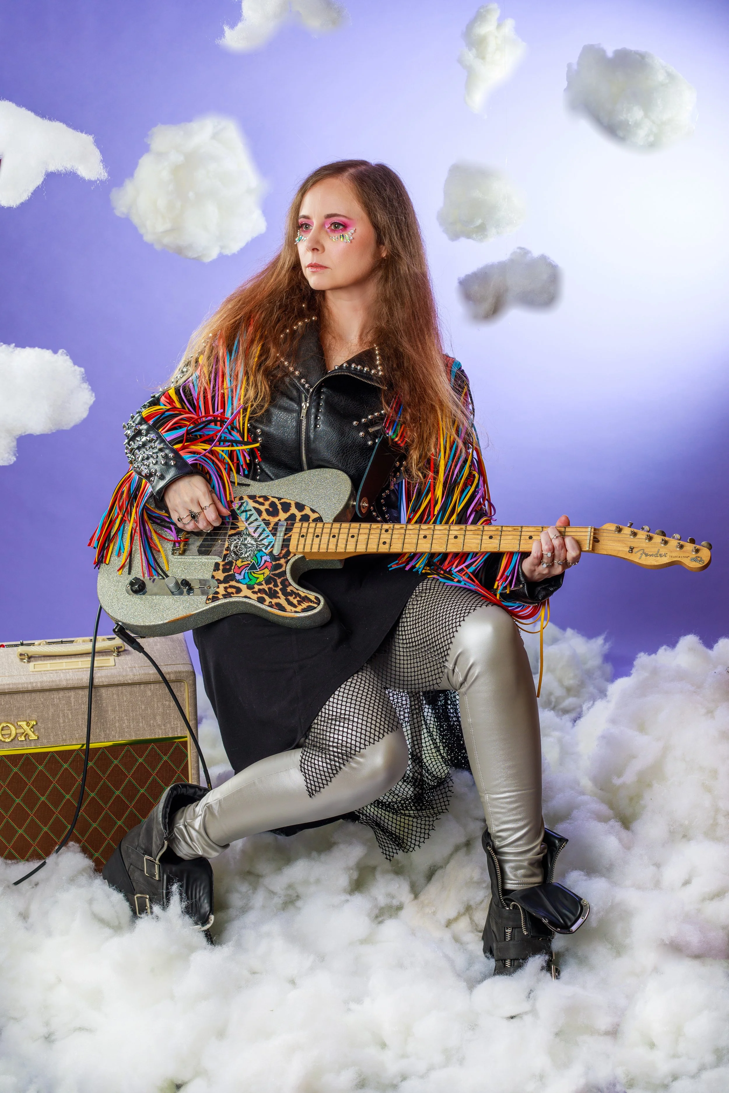A woman with long, wavy hair, wearing colorful makeup, a black leather jacket with rainbow fringes, metallic pants, and black boots, sitting on fluffy clouds with a purple background, holding an electric guitar with a leopard print and glittery finis