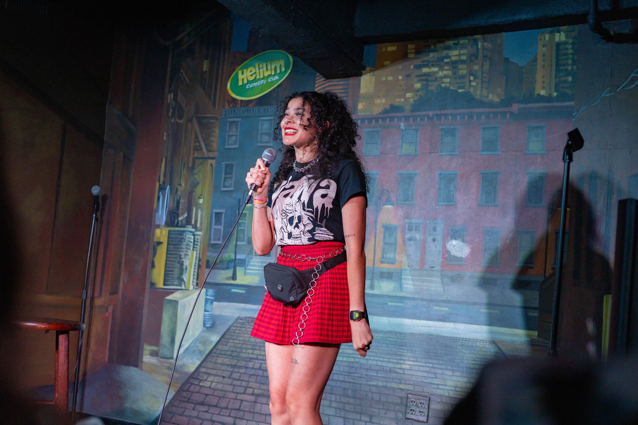 A woman with curly black hair and red lipstick stands on stage, smiling and holding a microphone. She is wearing a black graphic T-shirt, red plaid skirt, and a small crossbody bag. The background is a painted cityscape with a sign that says 'Helium comedy club.'