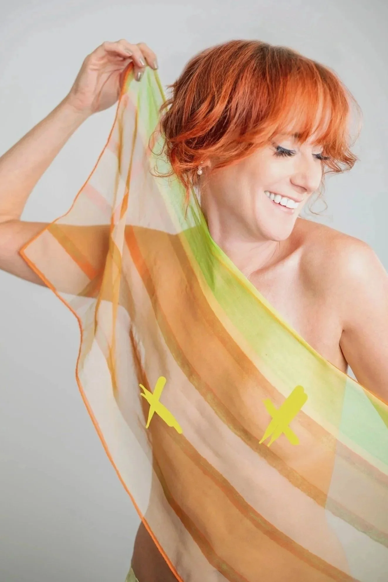 A woman with red hair smiling and holding a sheer, colorful scarf in front of her, partially draped over her shoulder.