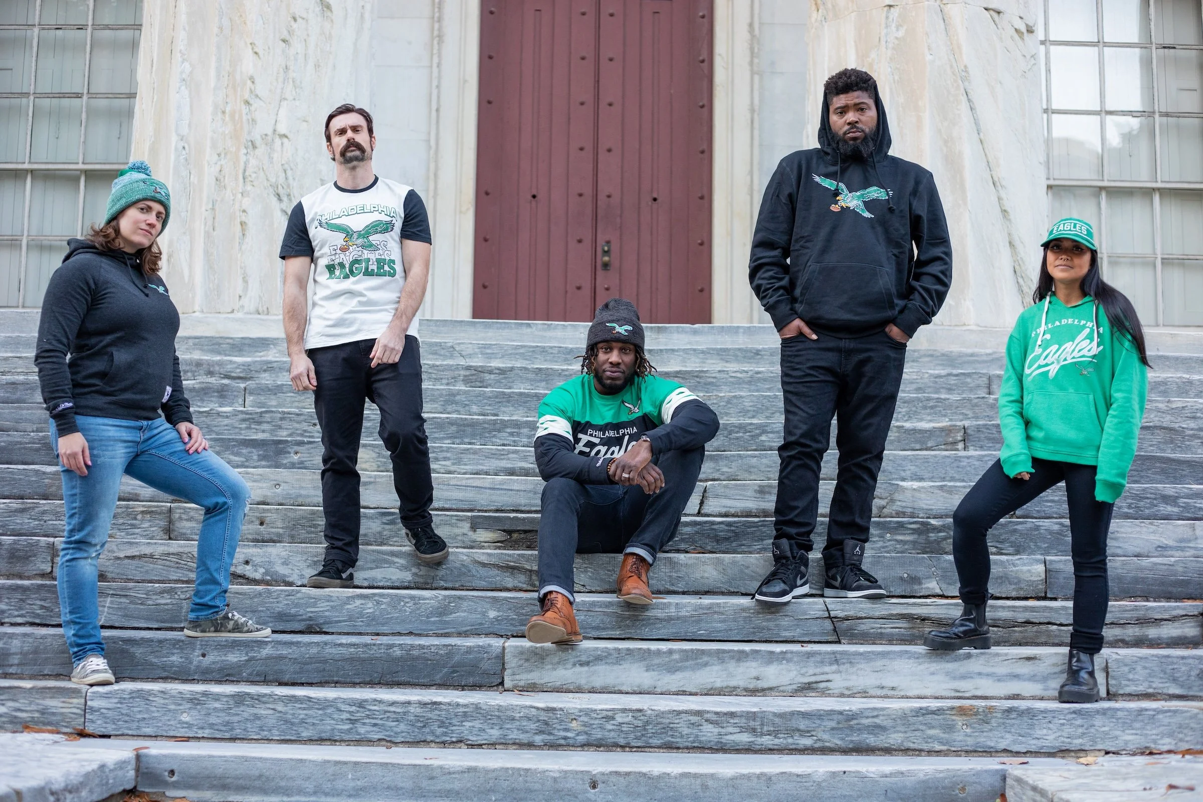 Group of five people standing and sitting on outdoor stairs in front of a building with large windows and a red door, wearing casual clothing and Philadelphia Eagles apparel.