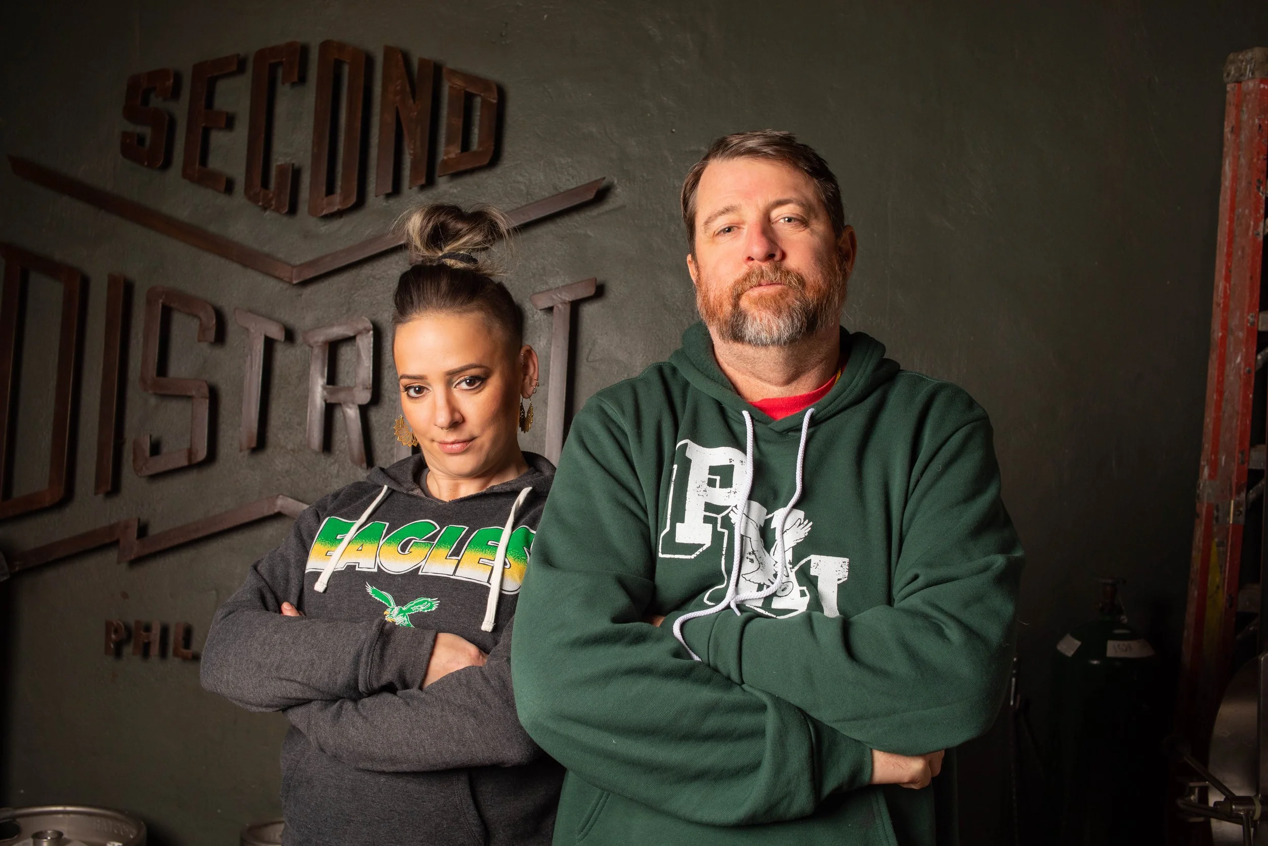 A man and a woman standing with arms crossed in a gym or storage room, with a sign on the wall behind them and equipment to the sides.