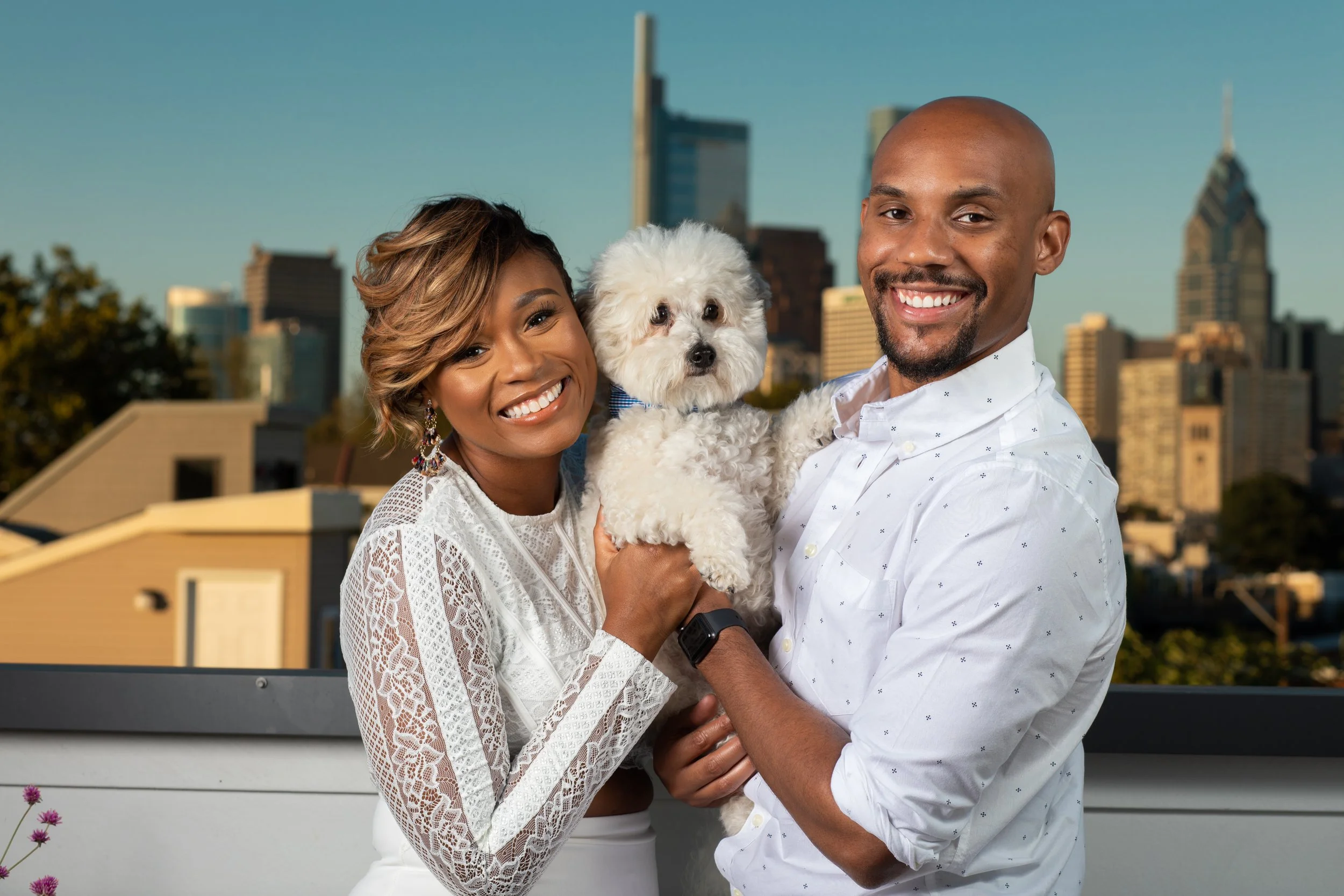 A smiling couple holding a small white dog with a city skyline in the background.