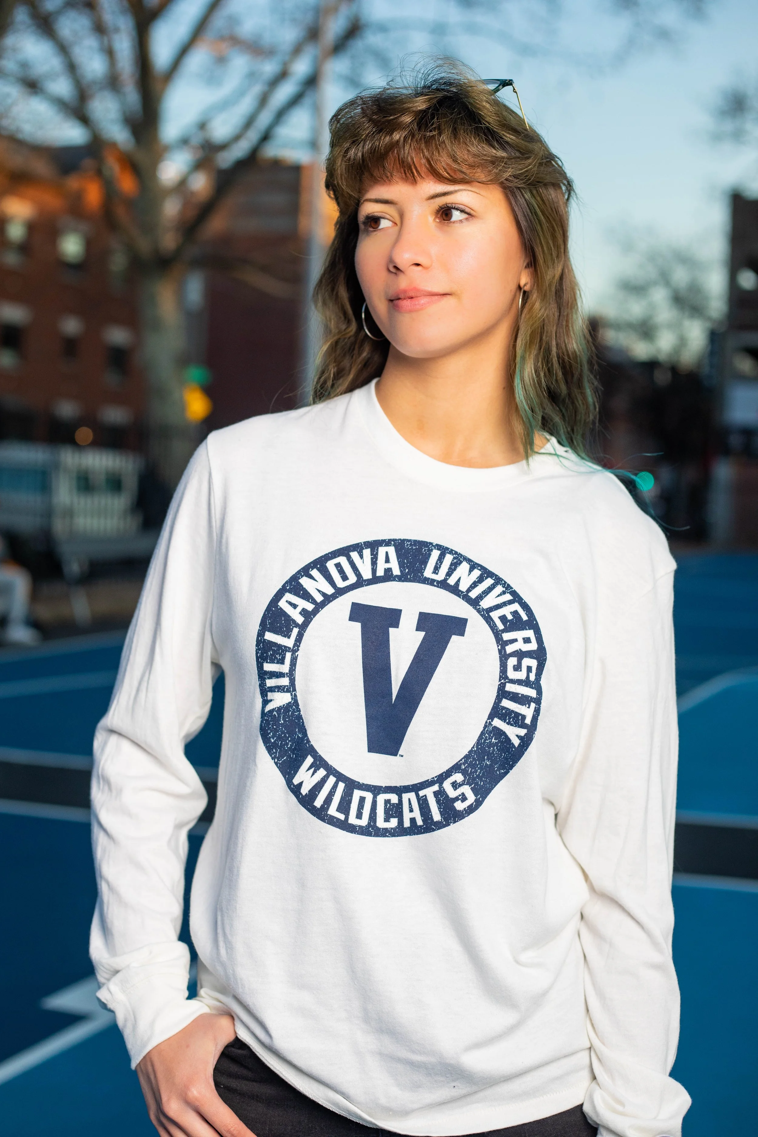 Young woman with brown hair and hoop earrings standing outdoors near tennis courts, wearing a white sweatshirt with 'Villanova University Wildcats' logo.