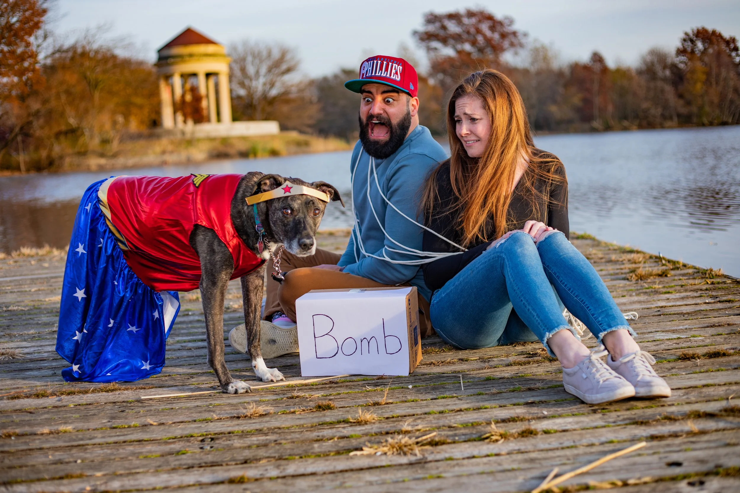 Two distressed people sitting on a dock next to a dog dressed as Wonder Woman, with a sign labeled 'Bomb', beside a lake with autumn trees in the background.