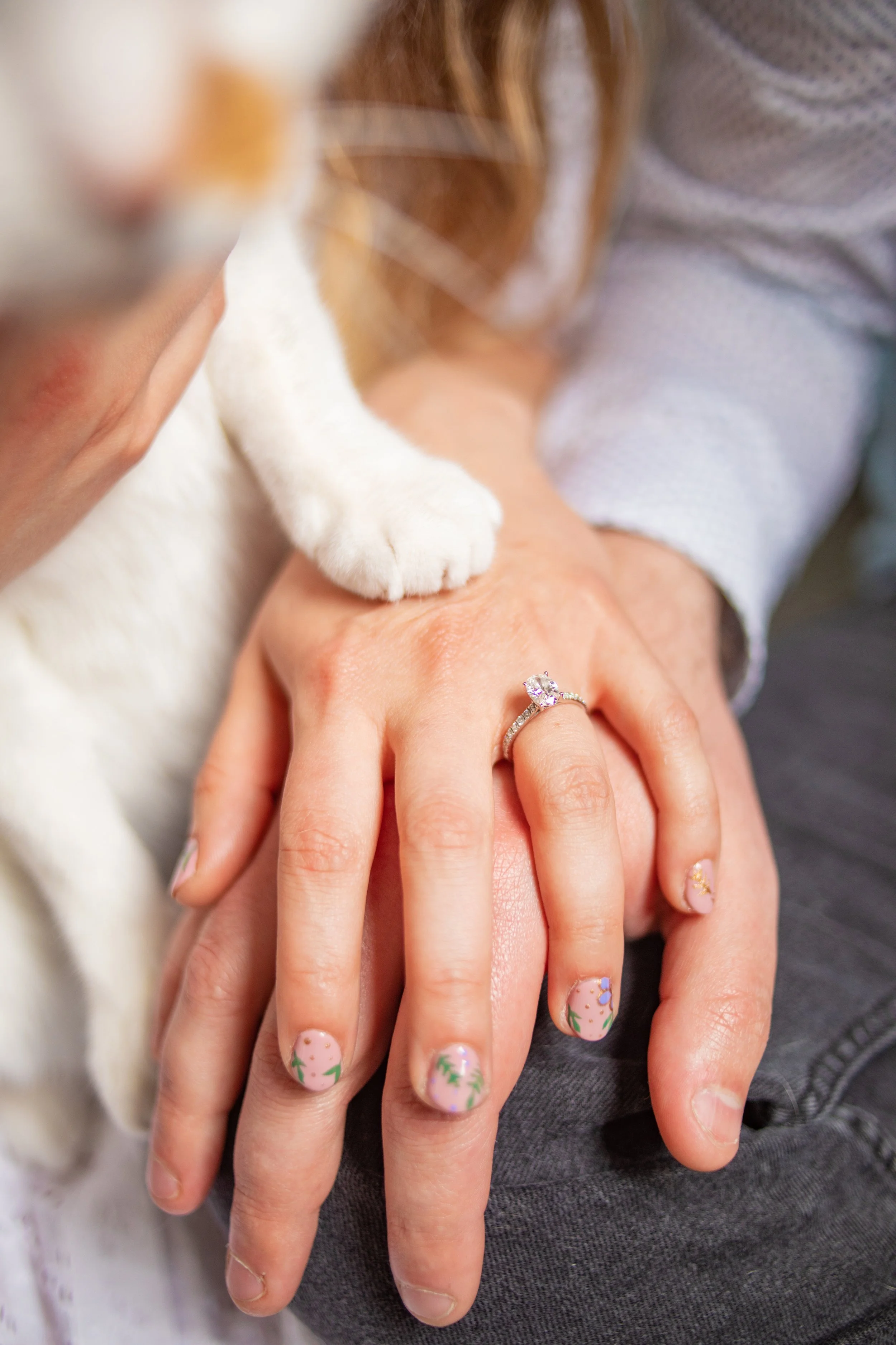 Close-up of a person's hand with a diamond engagement ring, resting on their lap, with a cat's paw resting on the hand. The person's nails have floral nail art.