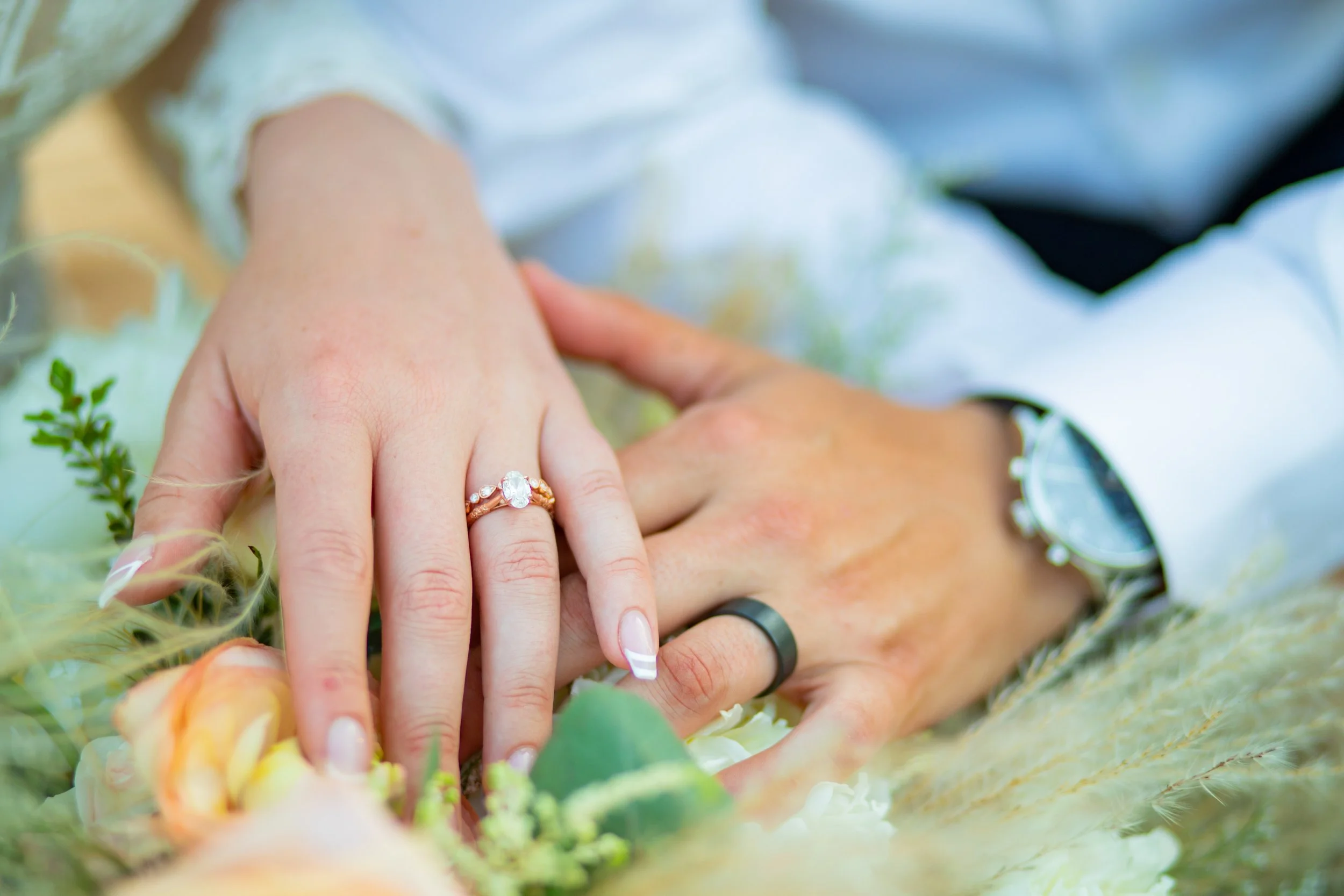 Close-up of a bride and groom's hands intertwined, showing wedding rings, surrounded by flowers and greenery.