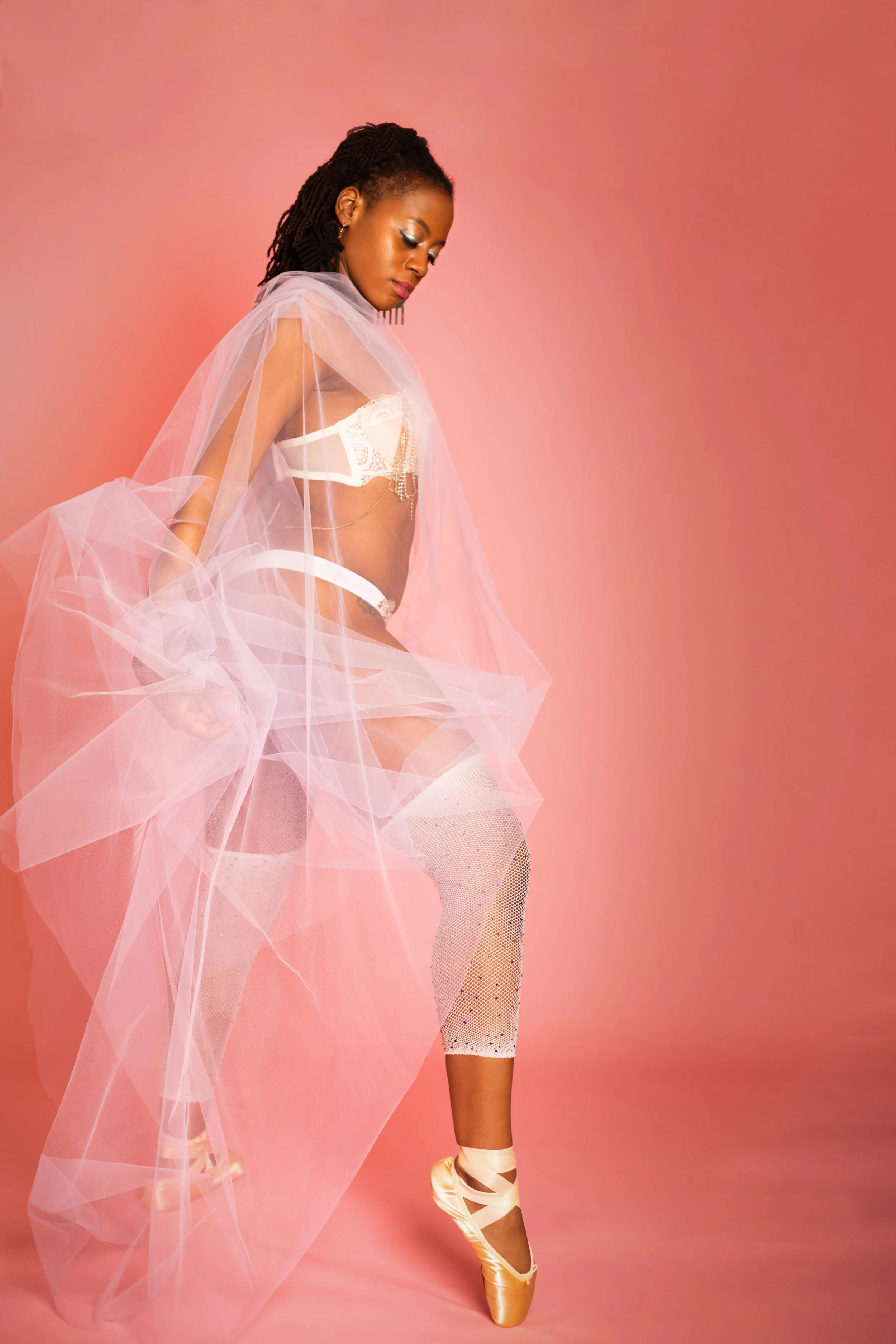 A female ballet dancer dressed in white lingerie, with a sheer pink cape, wearing pointe shoes, posing against a pink background.