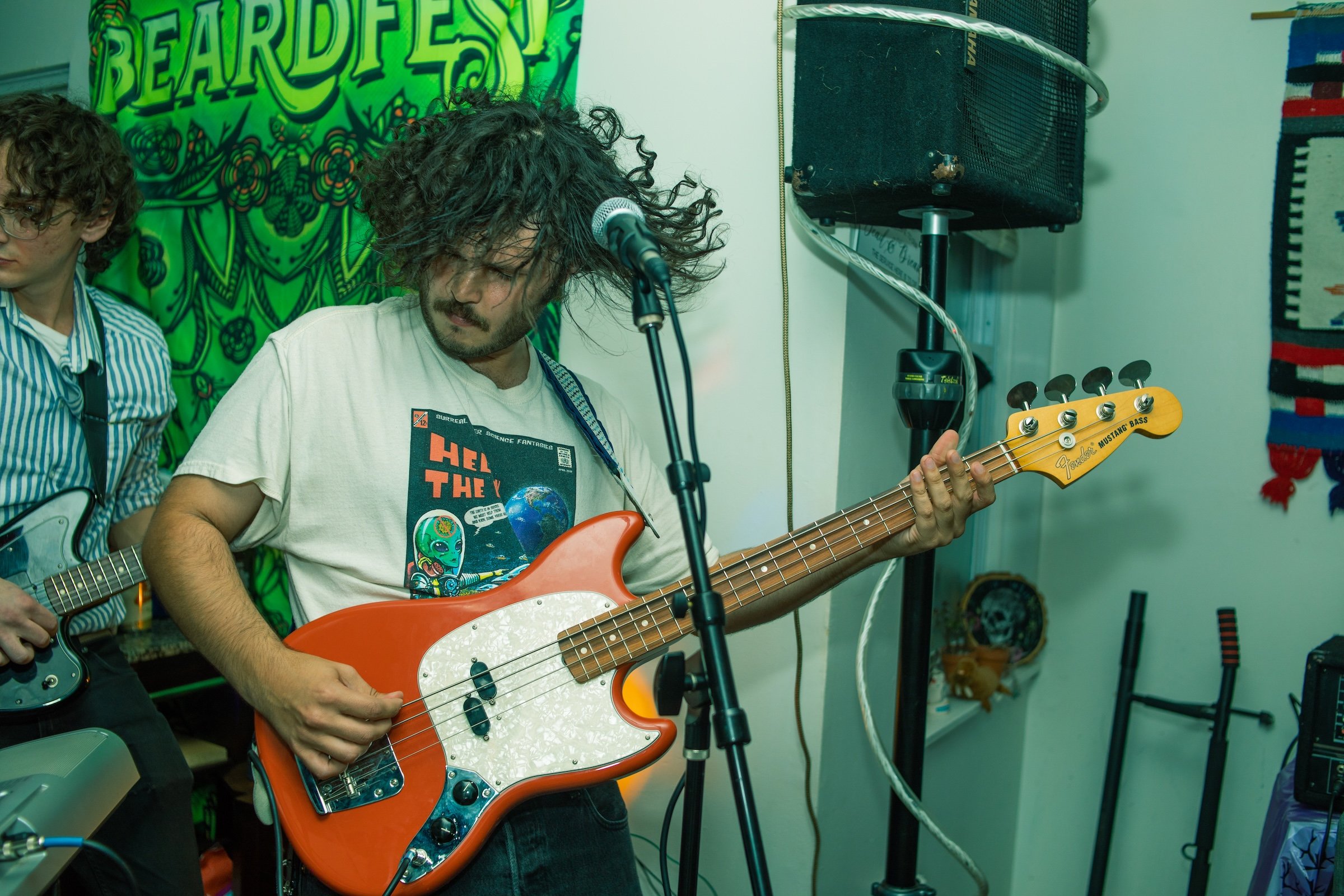 A man with curly hair and a beard playing a red electric bass guitar at a music event, with another person partially visible playing a guitar beside him, in a room decorated with green and patterned textiles.