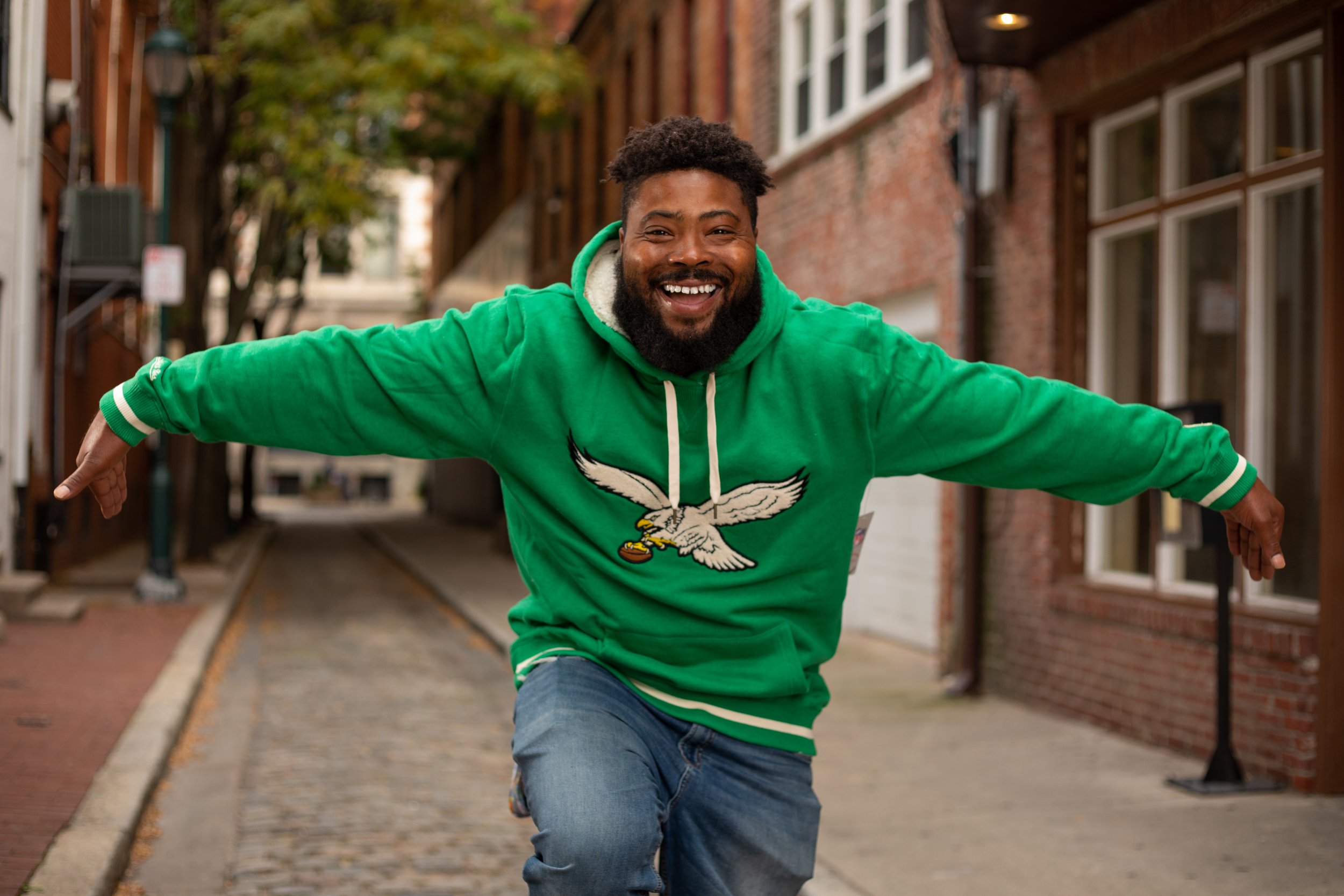 A man wearing a green hoodie with a sports team logo, smiling and stretching his arms out as he rides a skateboard on a city street.