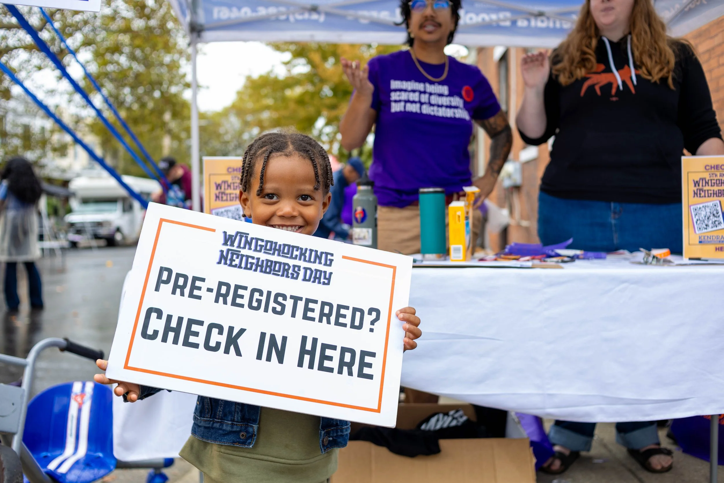 A young girl smiling and holding a sign that reads 'Windsocking Neighbors Day Pre-Registered? Check in Here' at an outdoor event with a table and two adults behind her.