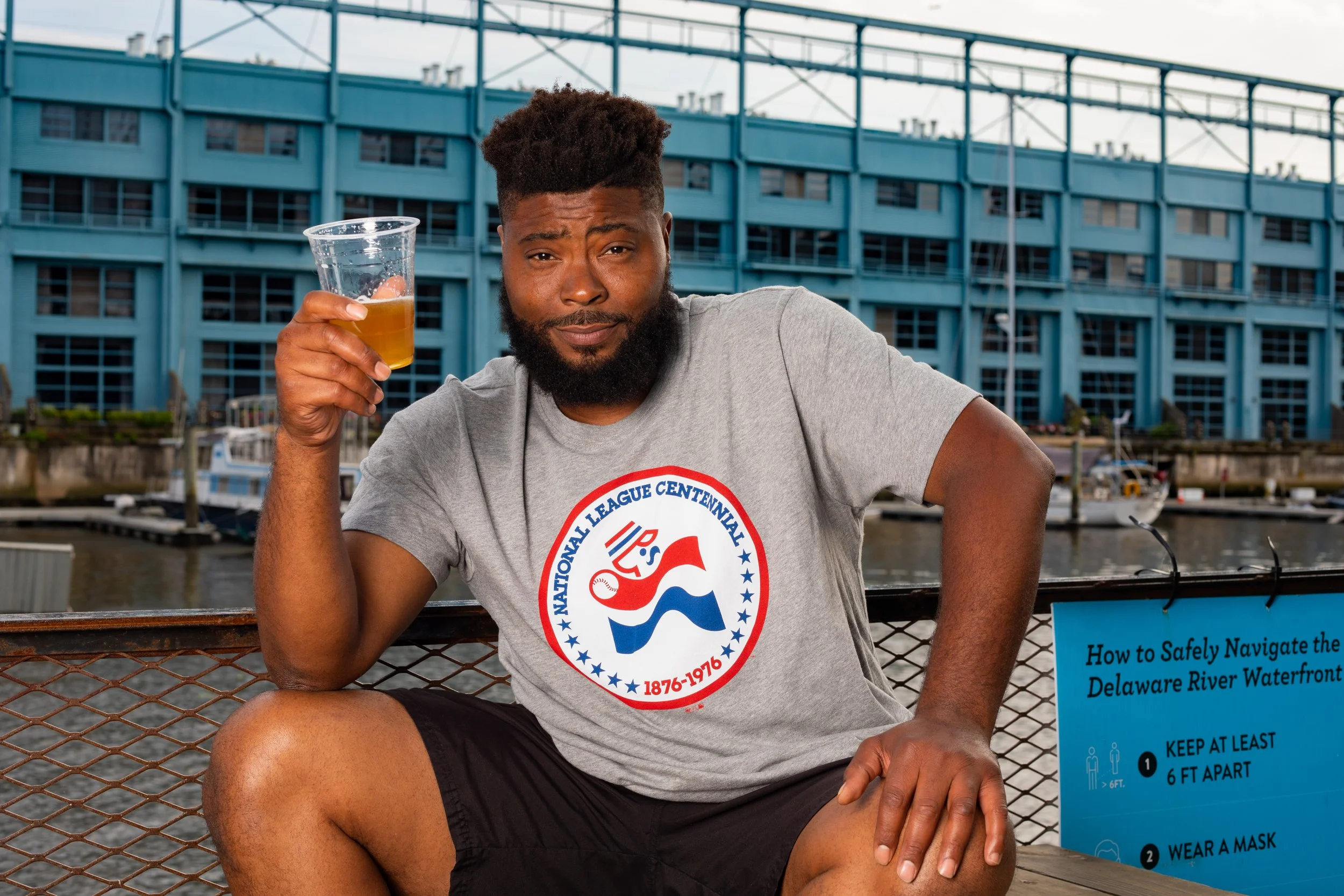 A man with a beard and short hair sitting by the waterfront, holding a plastic cup with a yellow beverage, wearing a gray T-shirt with a patriotic logo and black shorts.