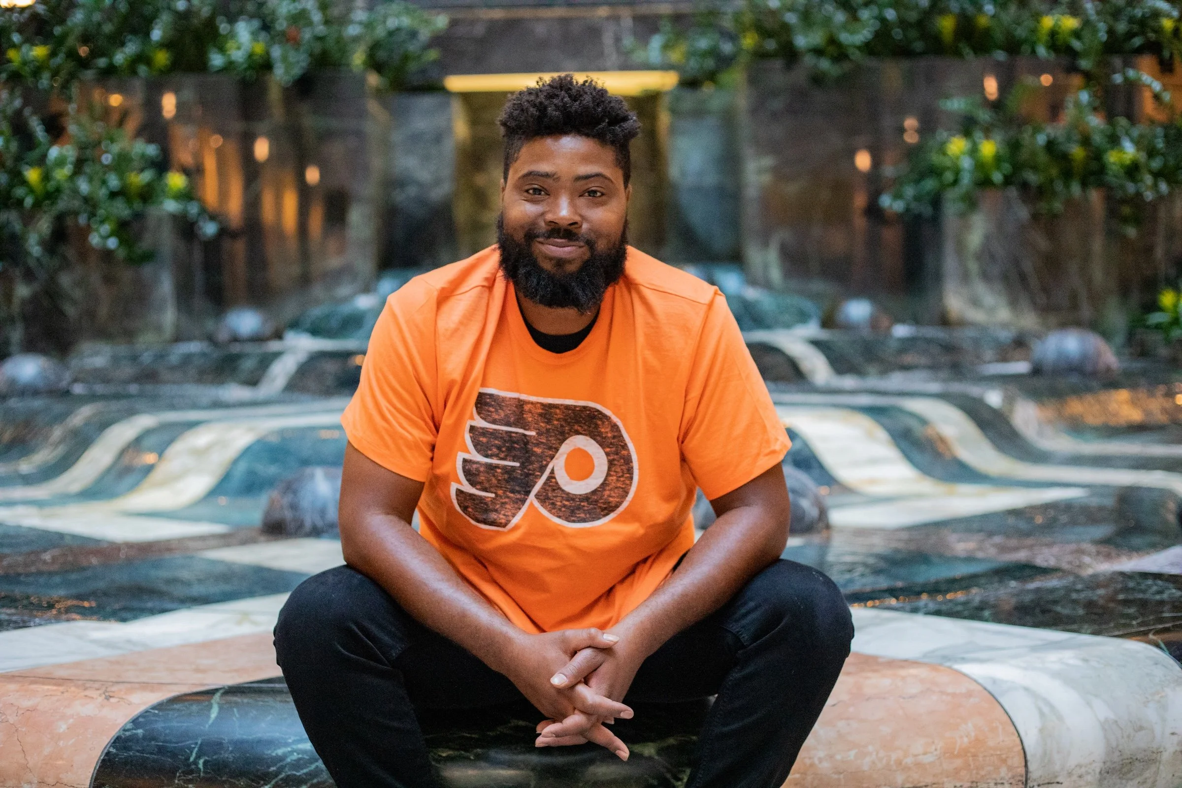 A man with a beard and curly hair sitting on a stone surface in front of a water feature, wearing an orange Philadelphia Flyers t-shirt and black pants.
