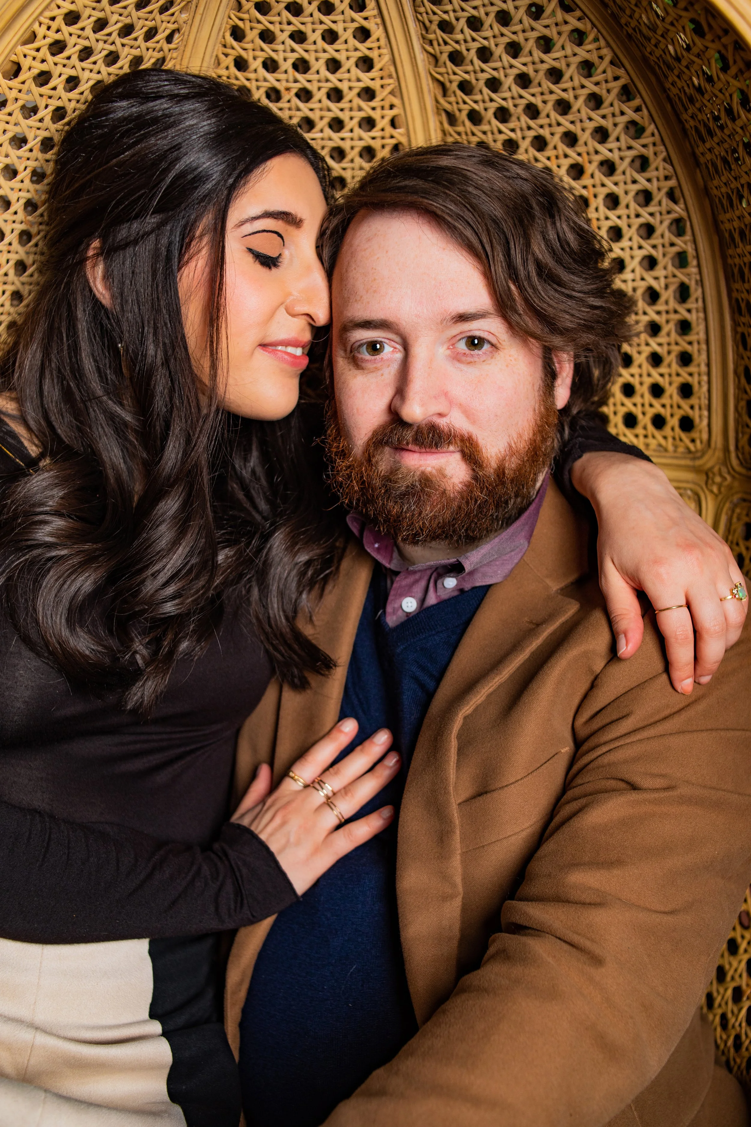 A woman with dark hair and a man with a beard are sitting close together, with the woman gently resting her hand on the man's chest. The woman has her eyes closed and appears to be whispering or about to kiss the man's temple, while the man looks dir