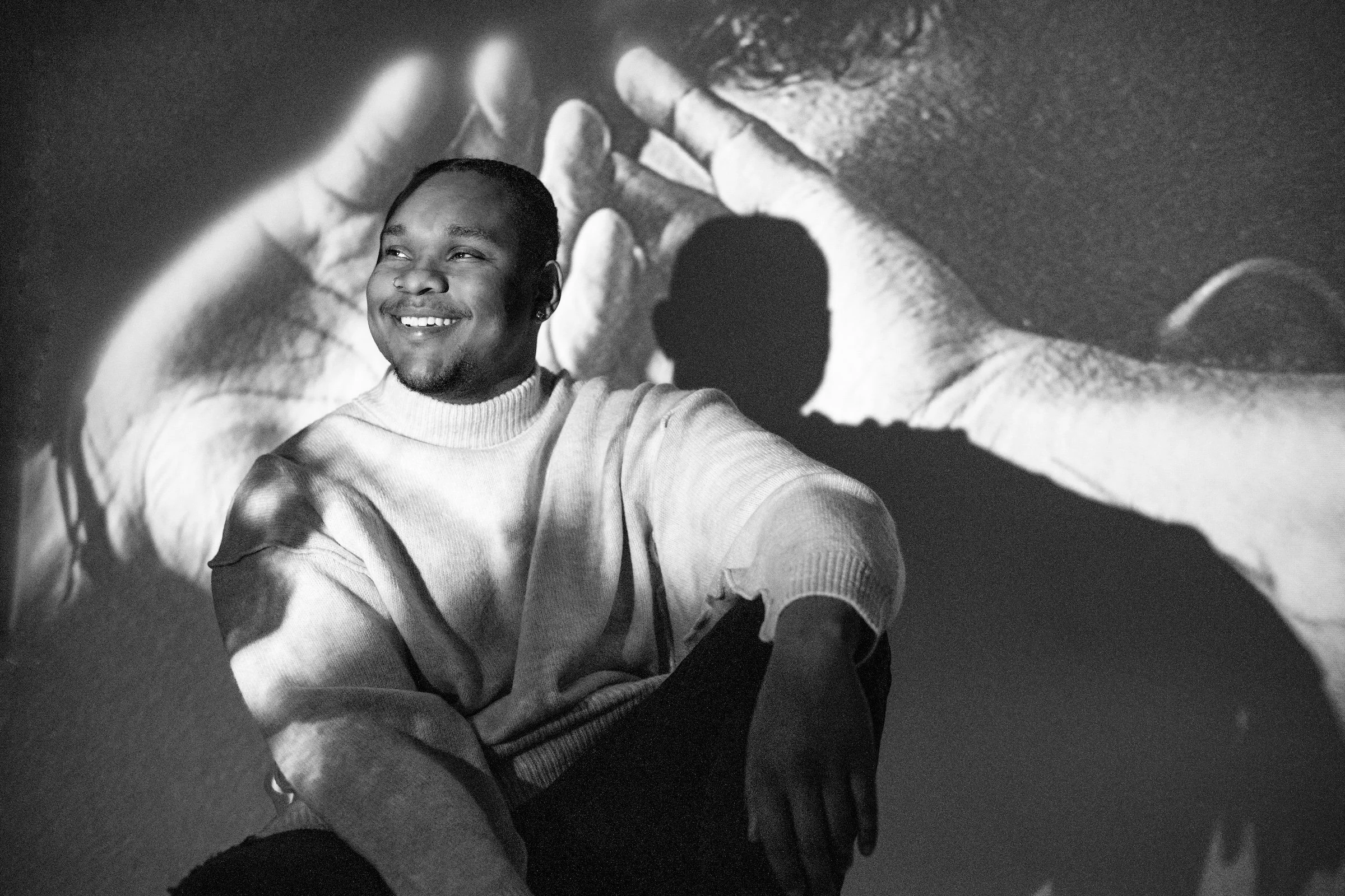 A young man sitting with a smile, wearing a light-colored sweater, with a shadow of his profile cast onto a large black-and-white photograph of hands in the background.