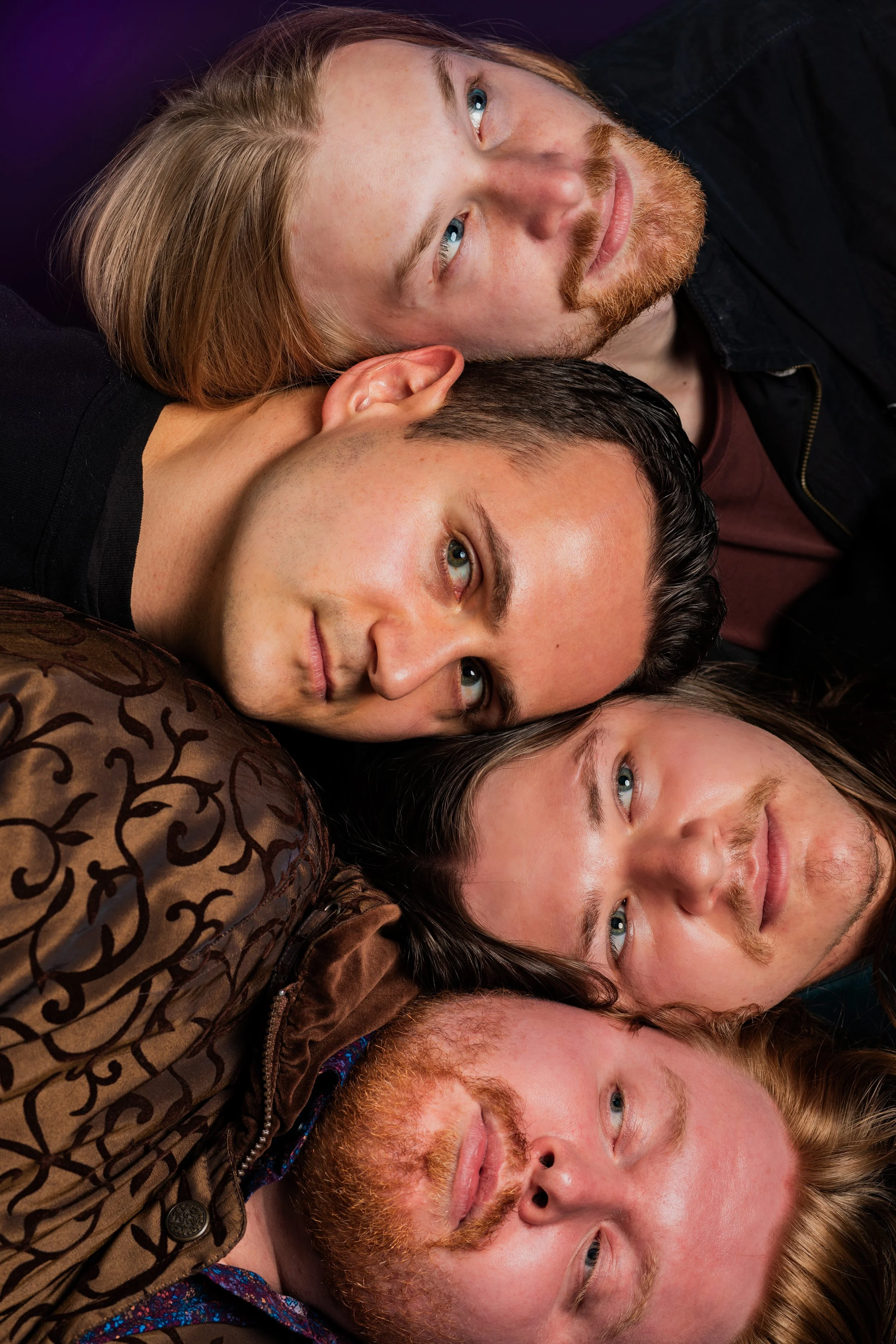 Close-up of five men lying on the floor, stacked closely with heads touching and faces visible, looking up.