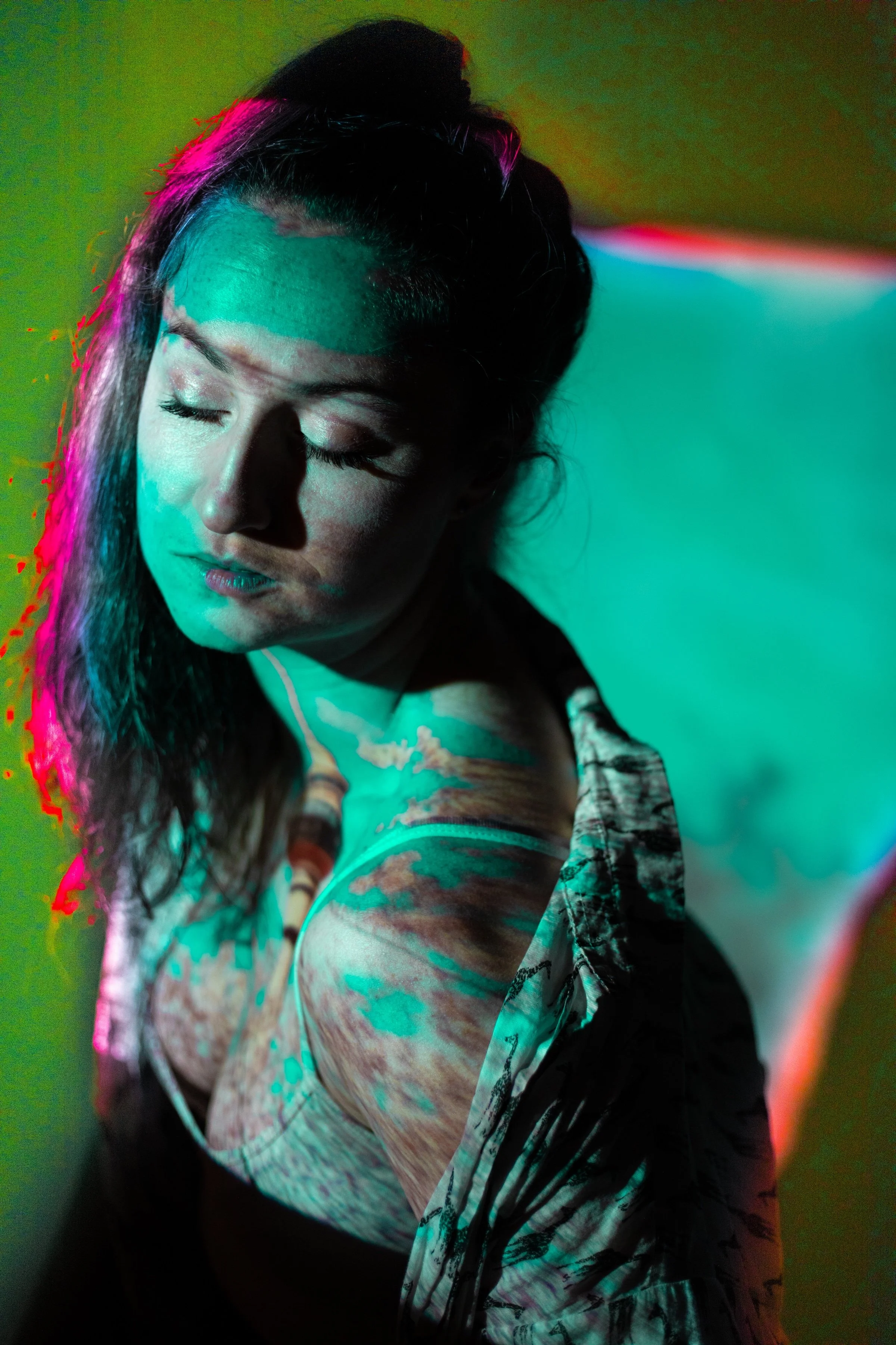 A woman with closed eyes and long hair illuminated by colorful lights, with a pattern projected onto her face and upper body.