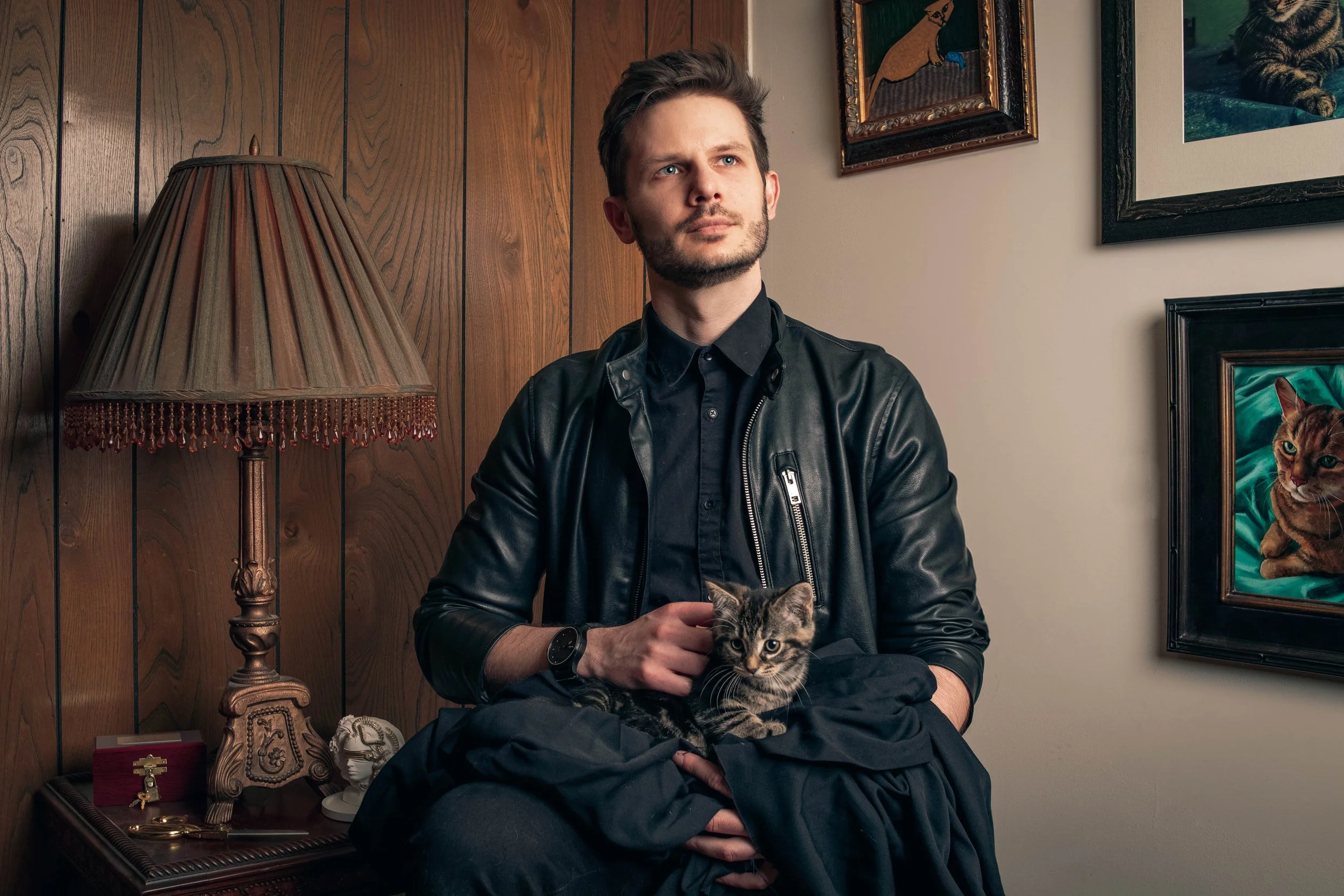 A man with light skin, short brown hair, and a beard, wearing a black leather jacket and black shirt, holds a tabby kitten on his lap in a room with dark wooden walls and framed artwork, standing next to a traditional lamp and jewelry box.