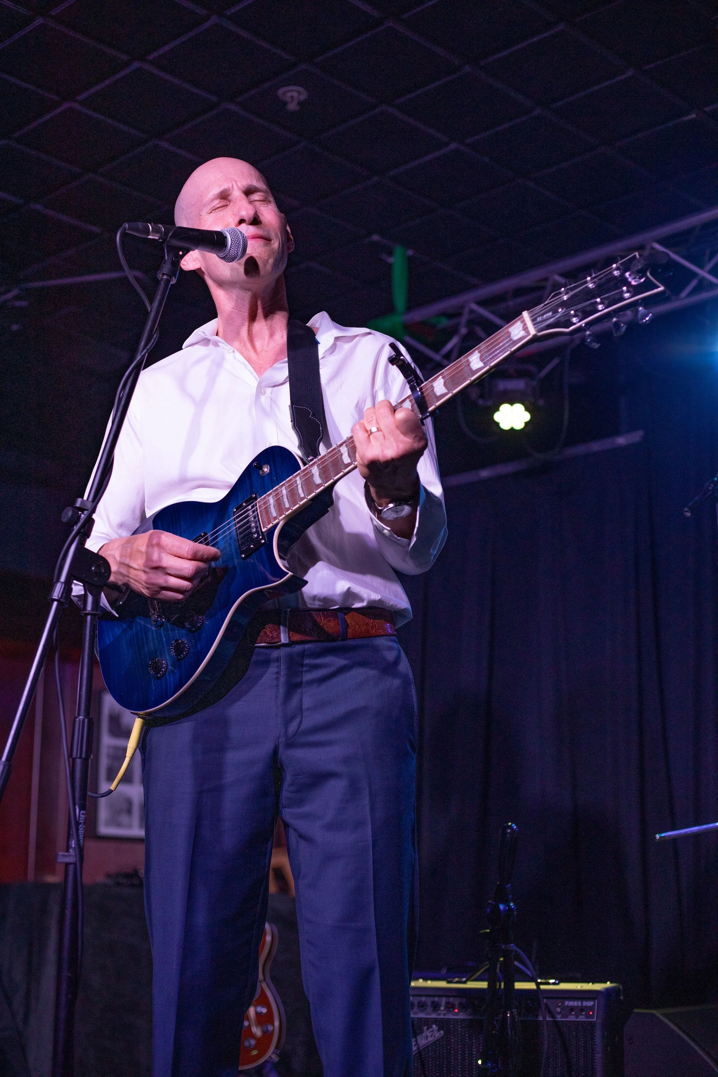 A man with a bald head singing with eyes closed and playing an electric guitar on stage, wearing a white shirt and dark pants, with stage lights in the background.