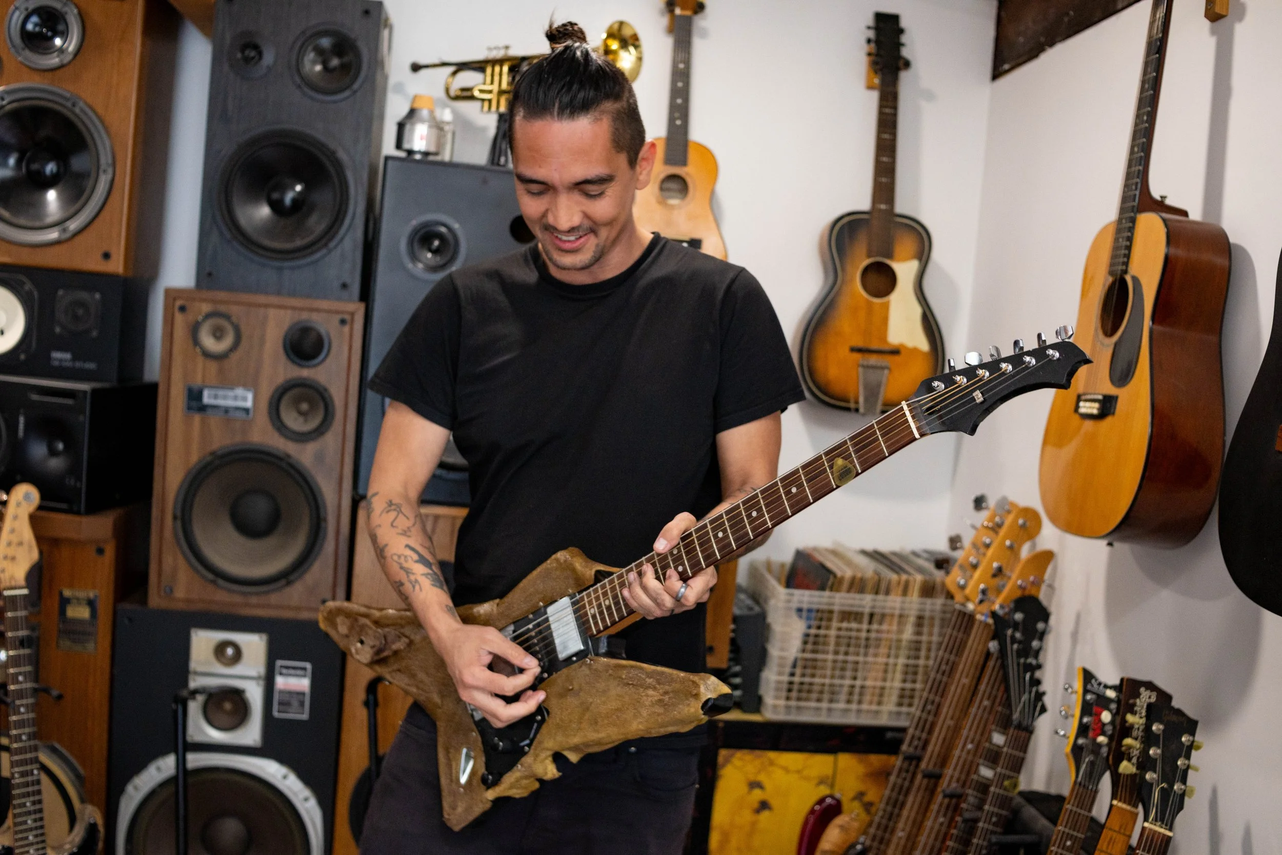 A man with a man bun hairstyle, wearing a black t-shirt, playing an electric guitar with a natural wood finish in a music room filled with various guitars and amplifiers on the walls and shelves.