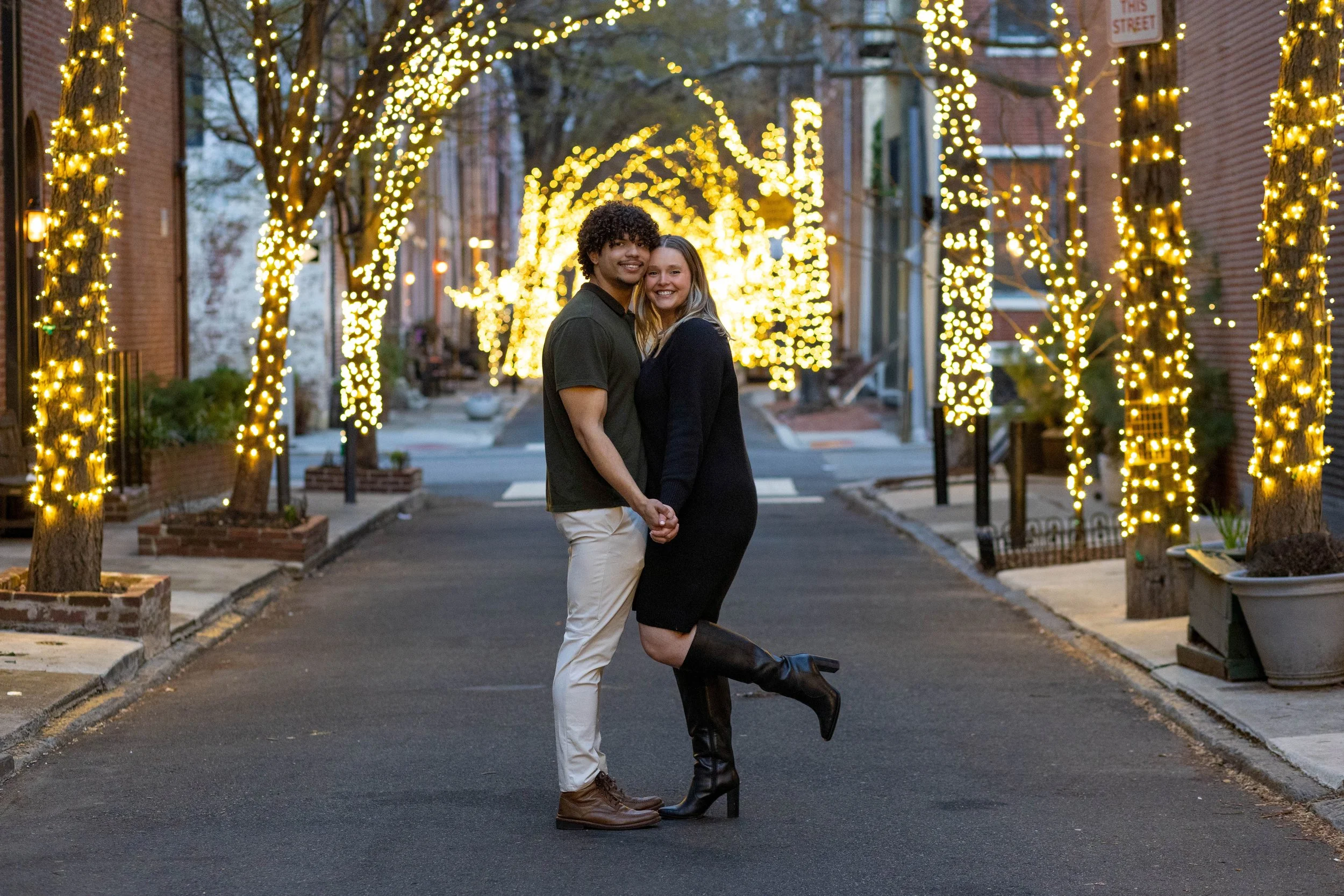 A couple is standing on a street decorated with yellow Christmas lights. They are holding hands, smiling, and facing the camera. The woman is wearing a black dress and high black boots, and the man is wearing a black shirt and white pants. The street