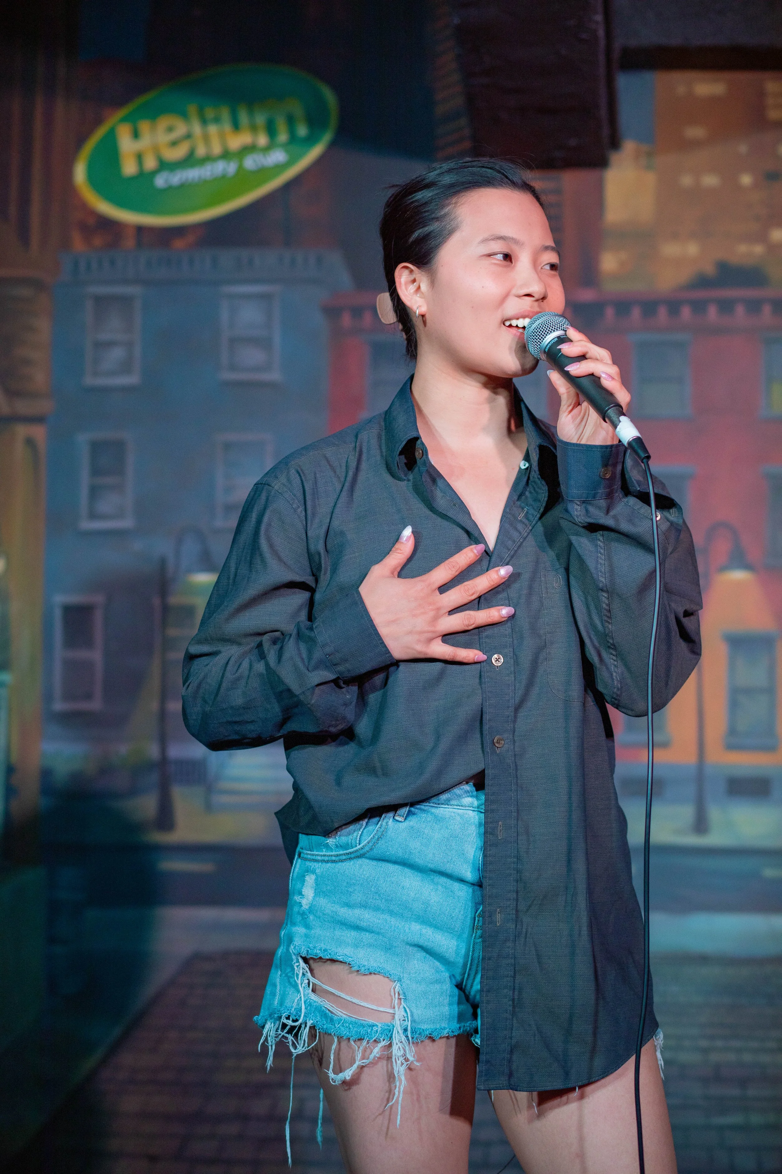 A woman standing on stage holding a microphone, wearing a black button-up shirt and ripped denim shorts, with a colorful cityscape backdrop behind her.