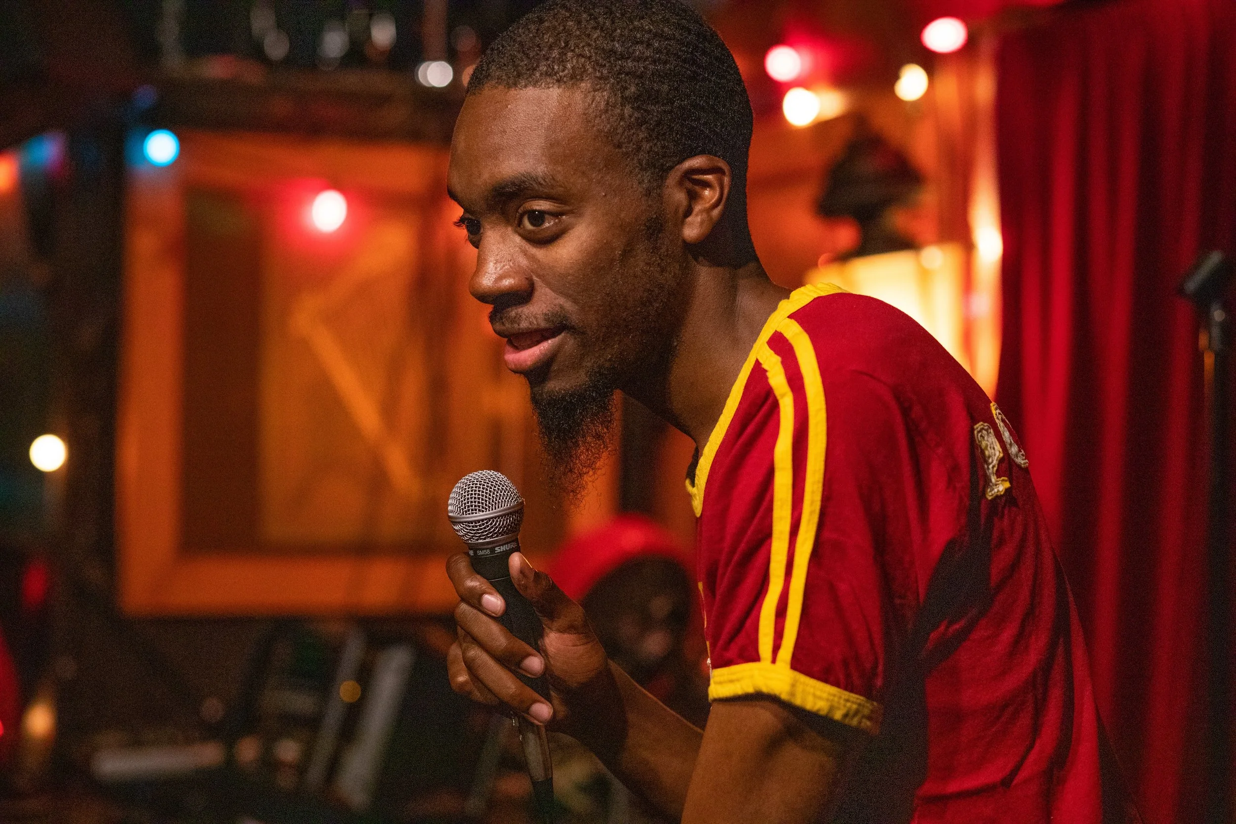 A man holding a microphone, wearing a red and yellow sports jersey, speaking or singing on stage in a dimly lit venue with colorful string lights and red curtains in the background.