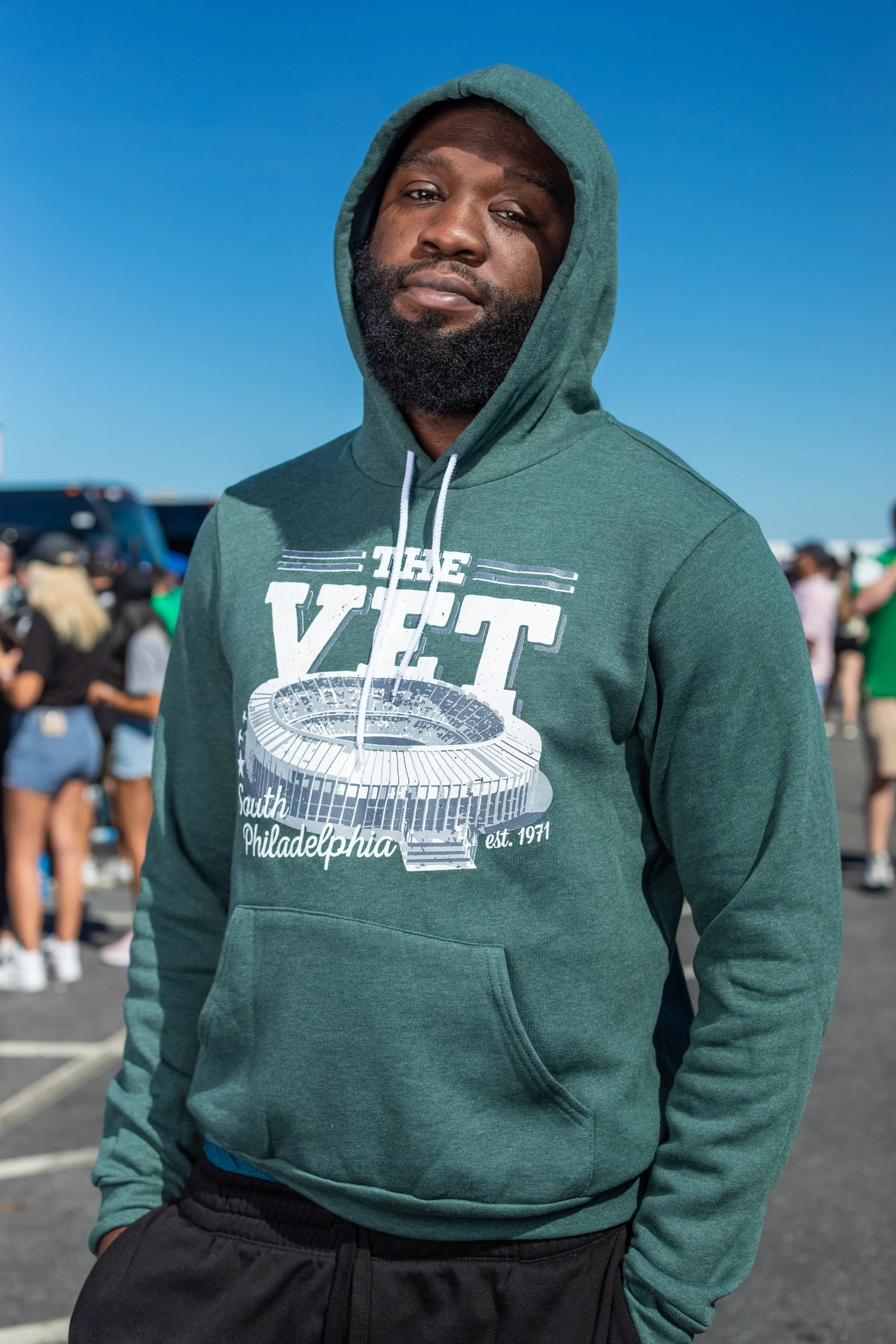 A man with a beard wearing a green hoodie with a stadium graphic and the words 'VET South Philadelphia est. 1971' stands outdoors on a sunny day, with a crowd of people in the background.