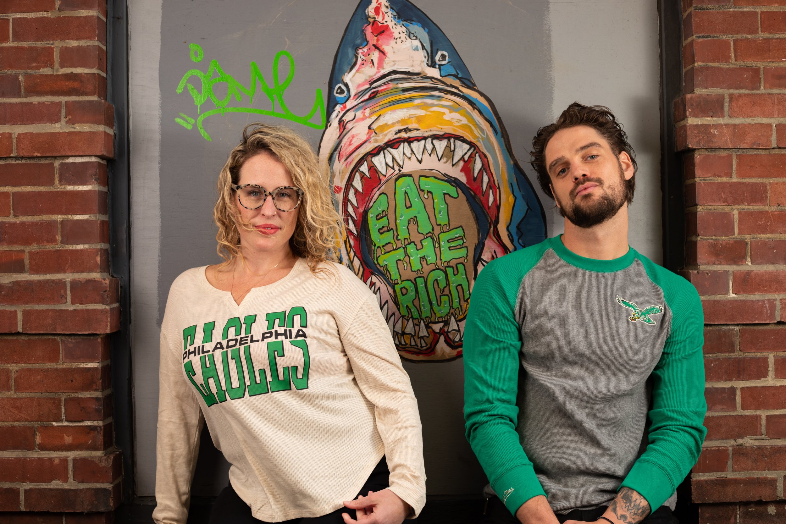 Two people standing in front of a mural featuring a shark with the words 'Eat the Rich' on its mouth. The woman on the left has curly blonde hair, glasses, and is wearing a beige shirt with green text that says 'I OWE TO PHILADELPHIA SHOULD'. The man