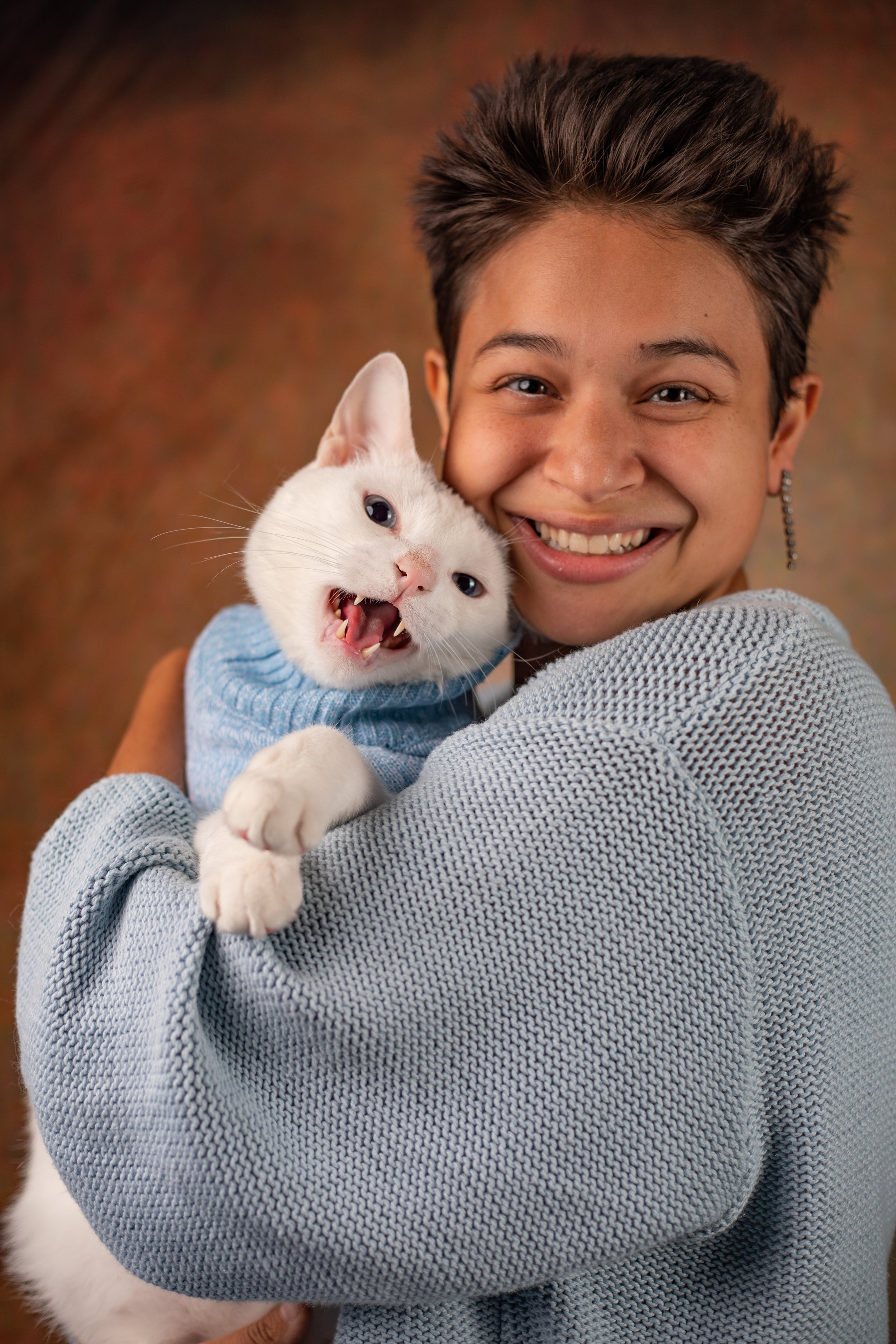 A person smiling while holding a white cat that is wearing a blue sweater and showing its teeth, in a warm-toned studio background.