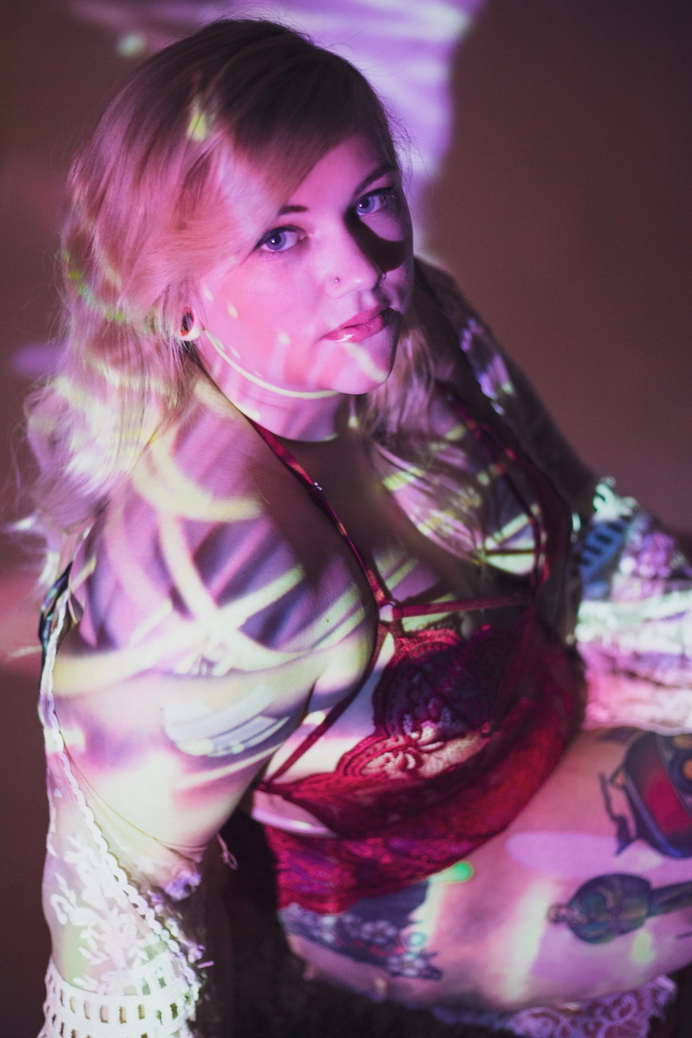 A woman with blond hair and blue eyes looking at the camera, illuminated with colorful lights and patterns.