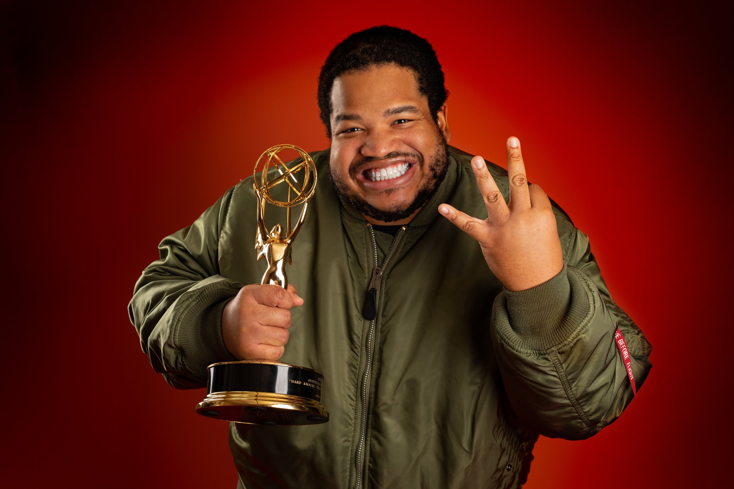 Man smiling and holding an Emmy award statue, making a victory sign with his fingers, against a red background.