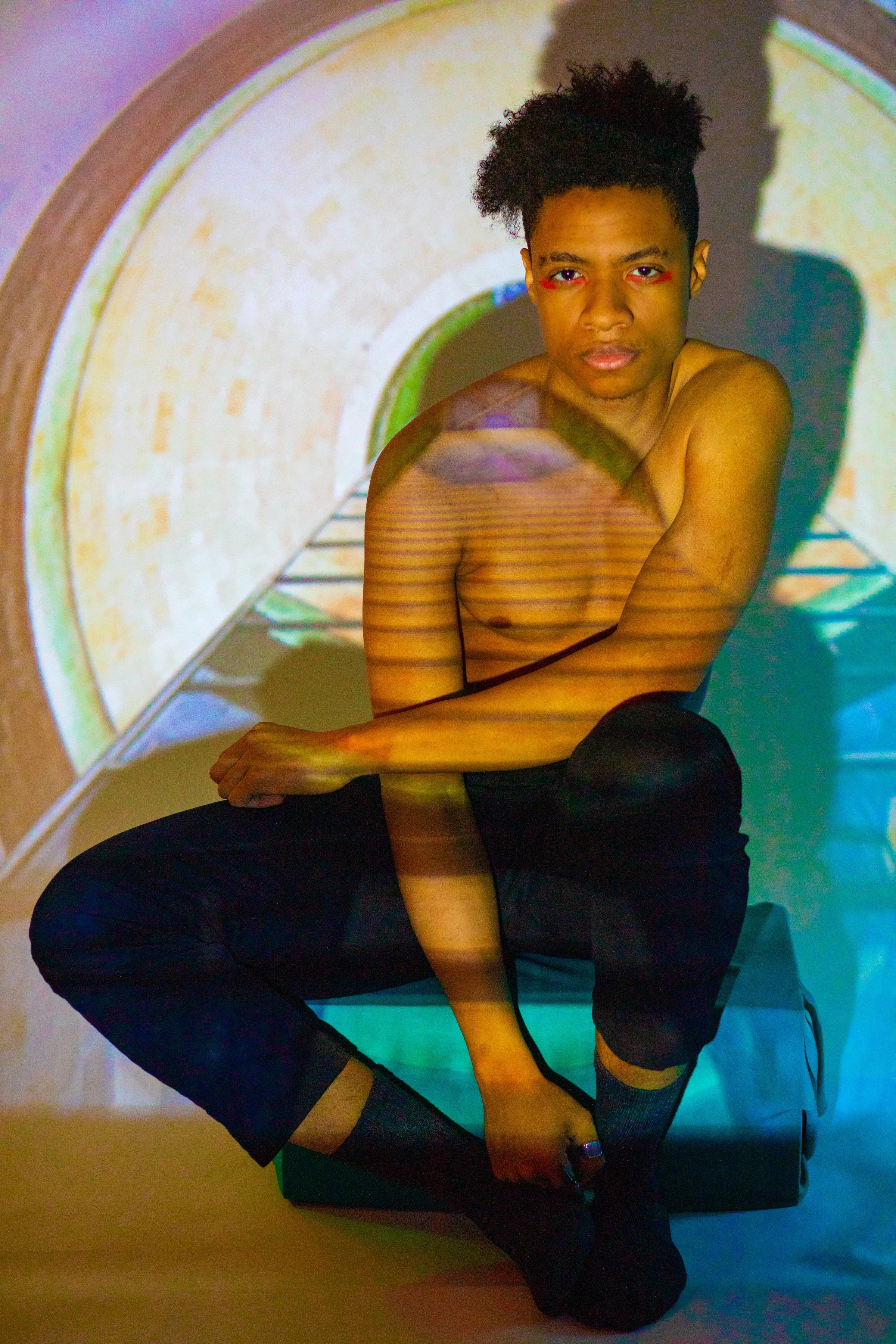 A shirtless young man with dark curly hair sitting with one knee up in front of a tunnel background projection, with rainbow-colored lines across his skin.