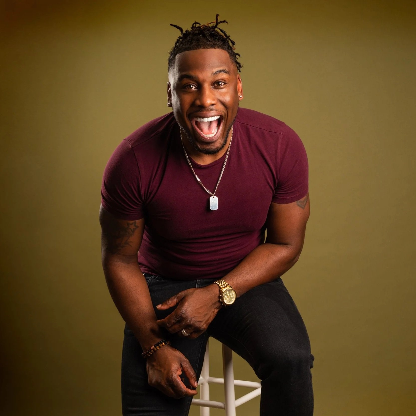 A man with dreadlocks, wearing a maroon t-shirt, gold watch, and necklace, sitting on a white stool with a surprised and happy expression against a plain olive background.