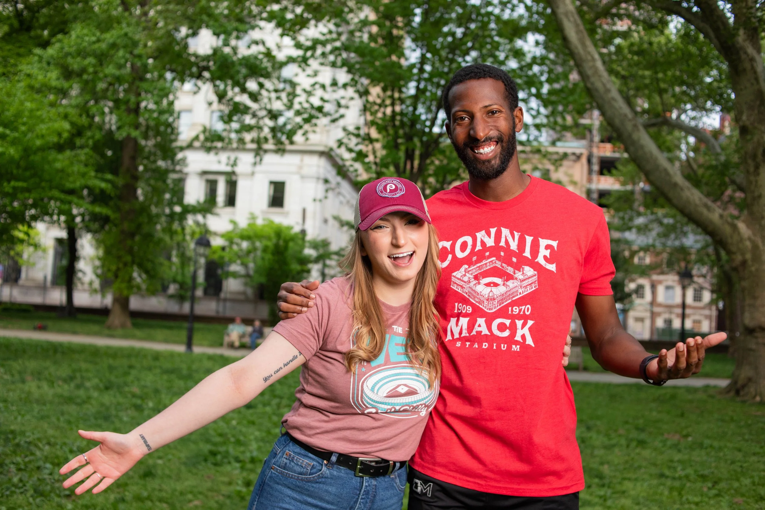 A woman and a man smiling and standing outdoors in a park with trees, grass, and buildings in the background. The woman is wearing a maroon baseball cap and a T-shirt, and the man is wearing a red T-shirt.