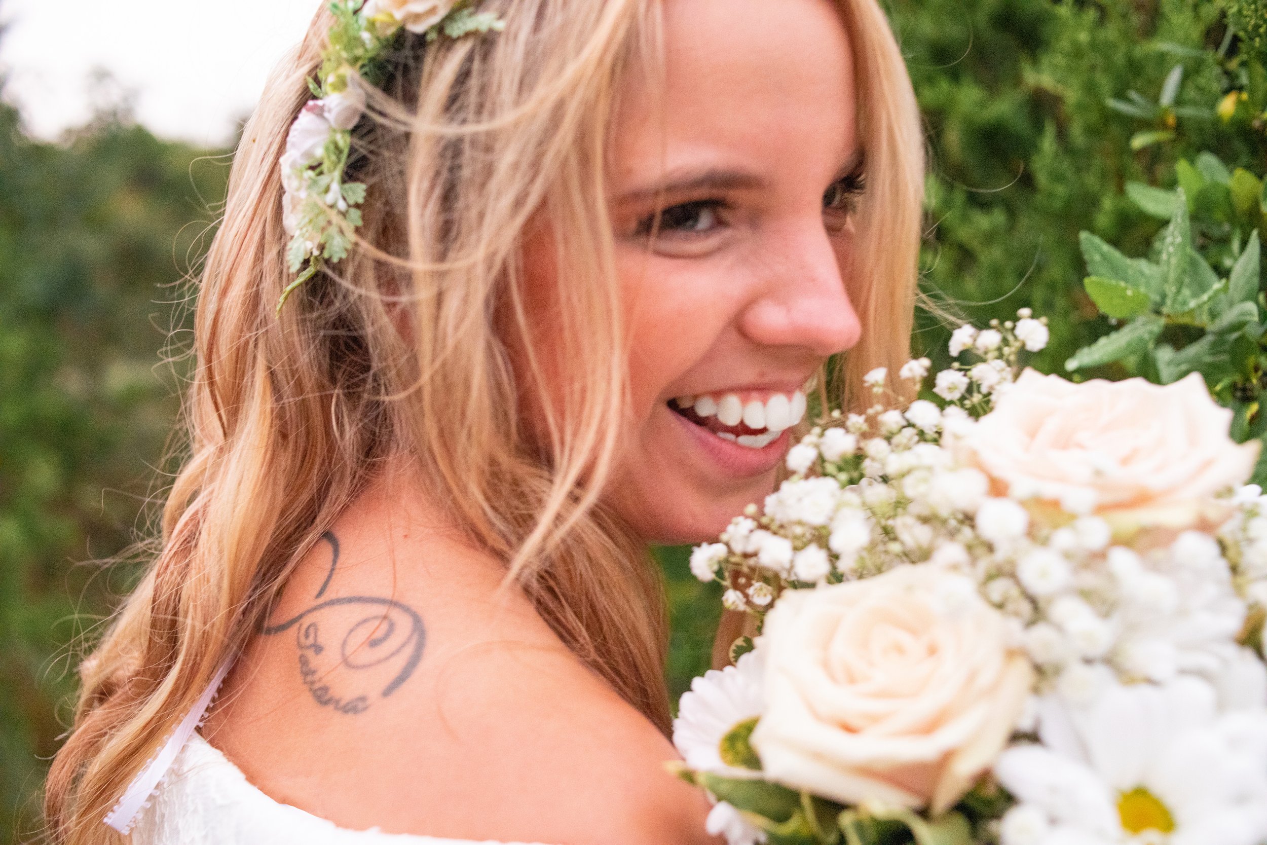 Smiling woman with blonde hair holding a bouquet of flowers, wearing a floral headpiece, outdoors with greenery in the background.