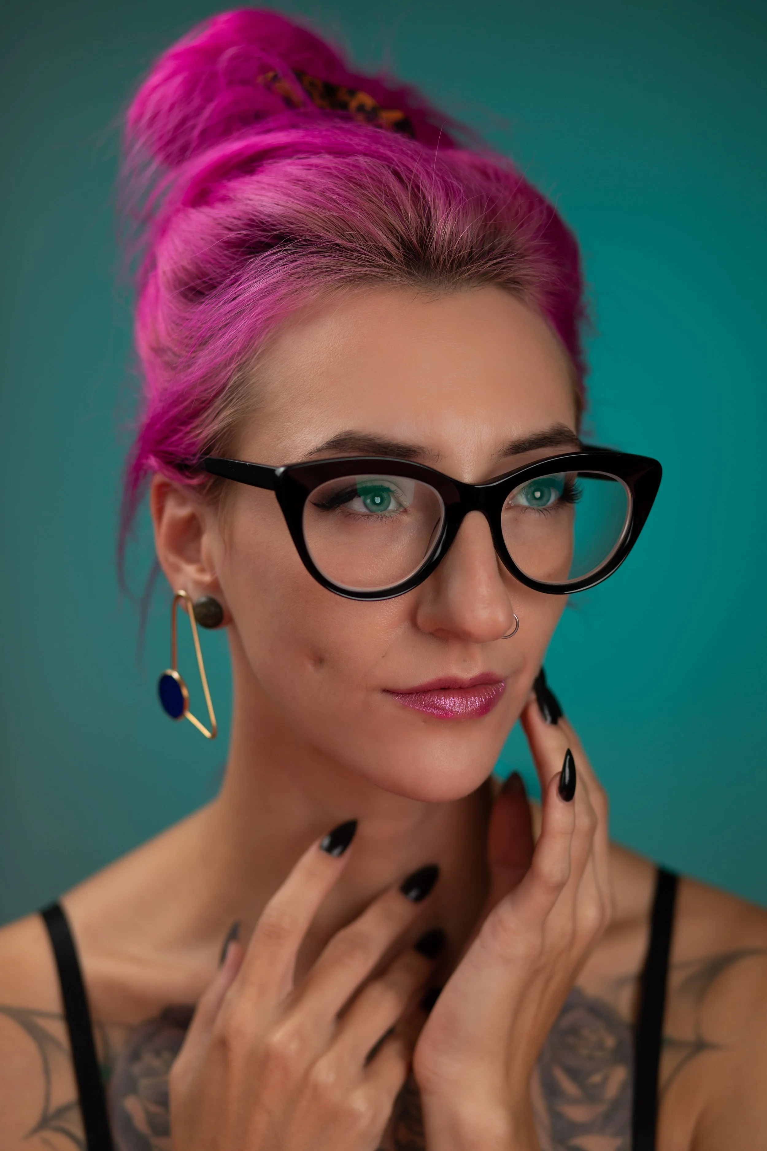 Portrait of a woman with pink hair styled in an updo, wearing black glasses, hoop earrings, and a black spaghetti strap top, posing against a teal background.