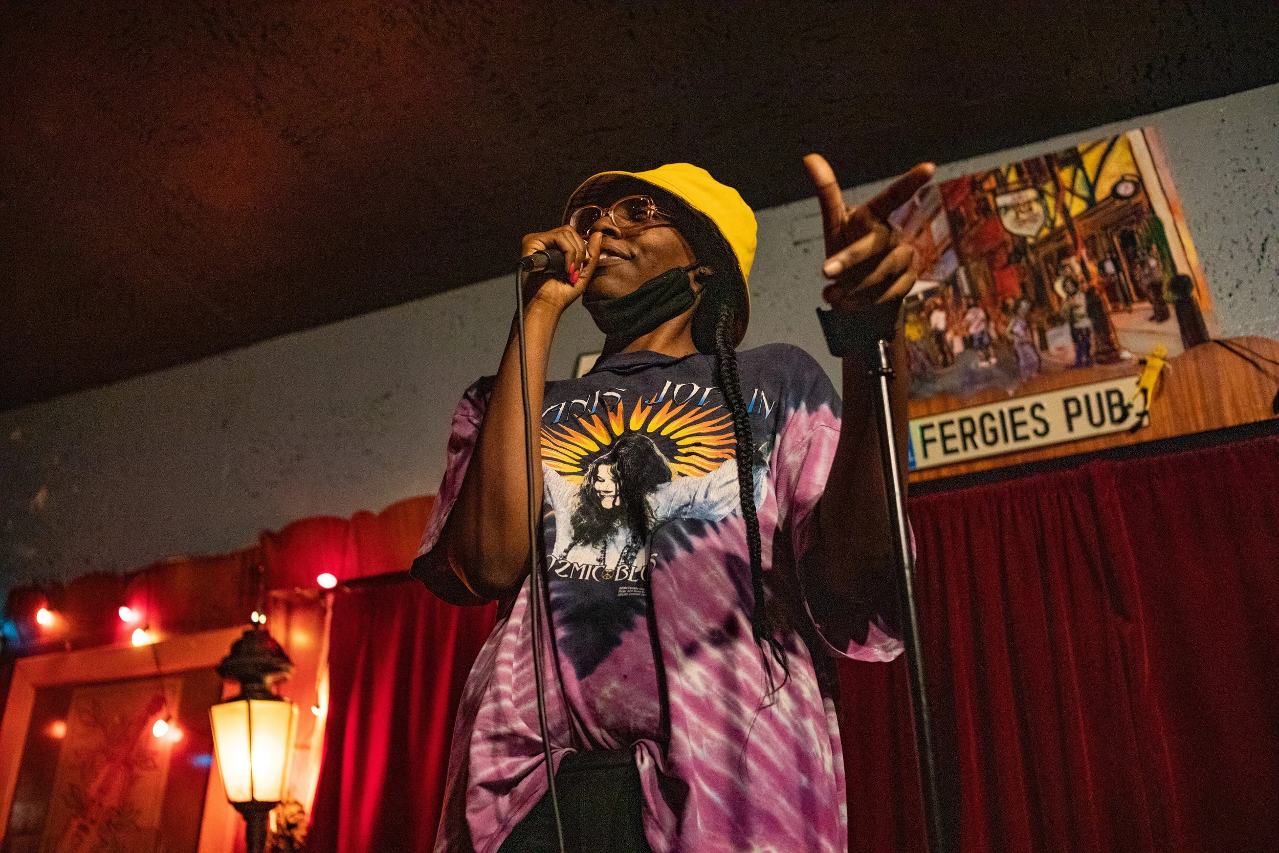 A woman with glasses, wearing a yellow beanie and a tie-dye shirt with a graphic of a woman, is performing with a microphone on a stage with red curtains and warm lighting.