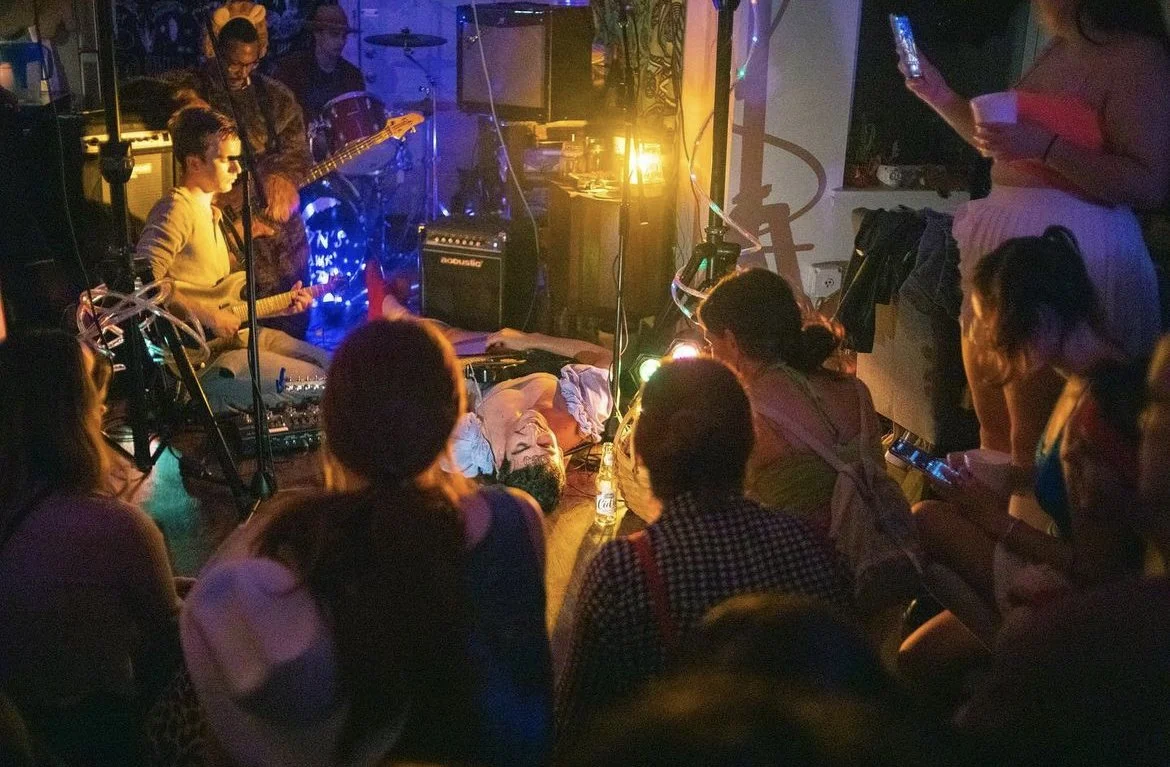 A live music performance with a singer lying on the floor surrounded by a small audience, some sitting on the floor and some standing, in a dimly lit venue with colorful lighting.