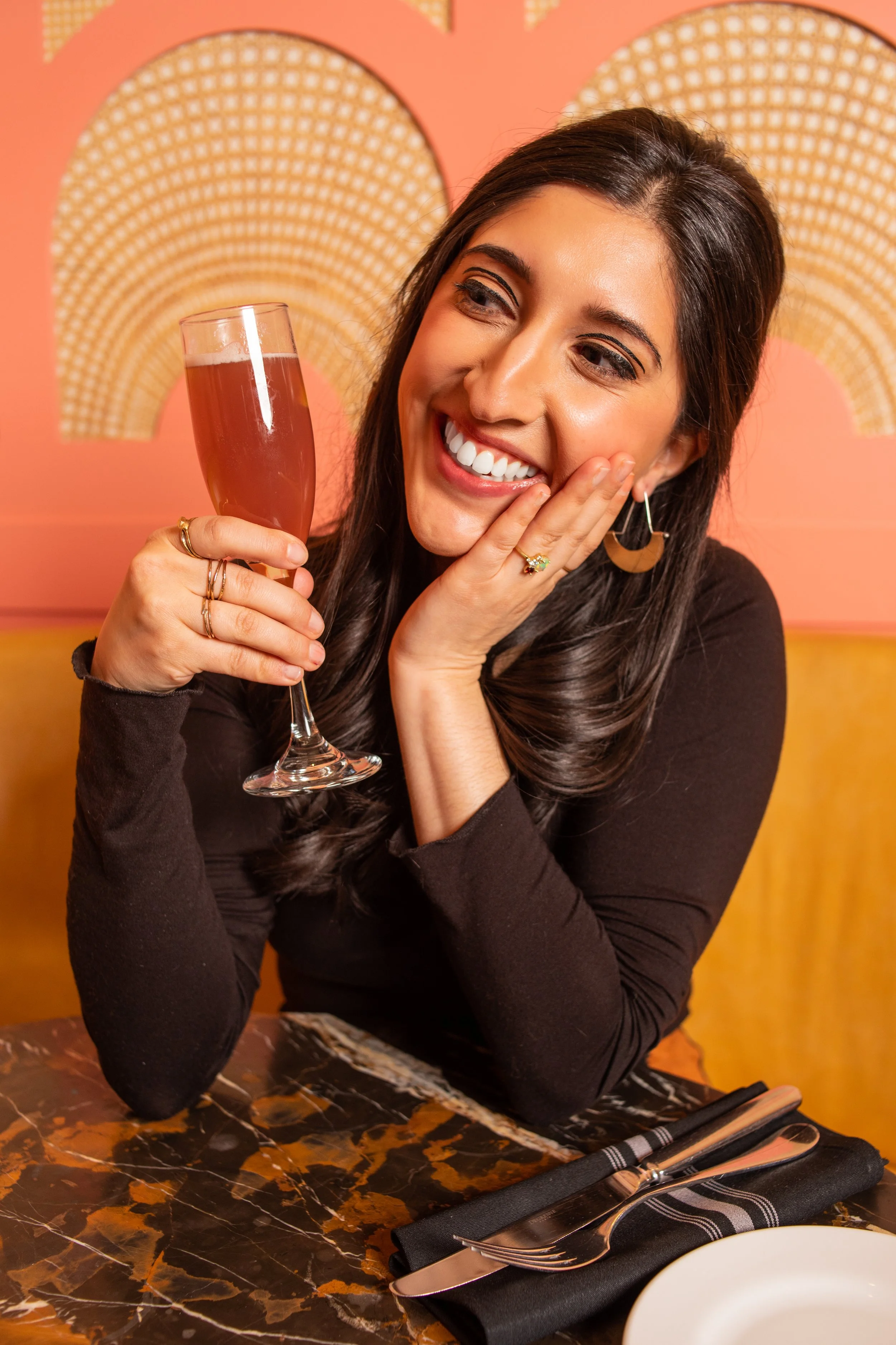 A woman with long dark hair, smiling, holding a glass of pink champagne, sitting at a table with silverware wrapped in a black napkin, in a restaurant with pink and yellow decor.