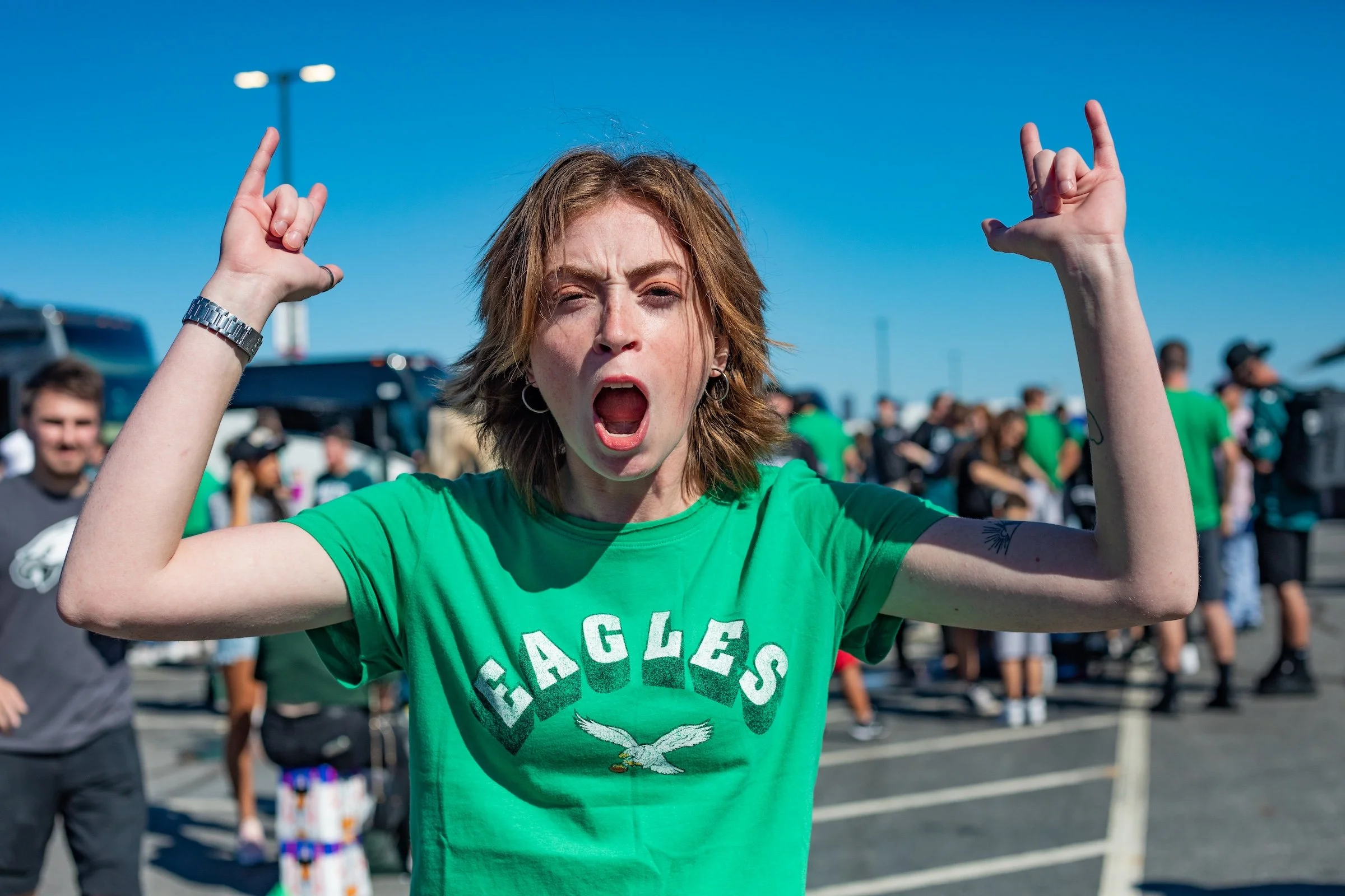 A woman with short brown hair, wearing a green Eagles T-shirt, making a rock hand gesture and shouting, at an outdoor event with a crowd in the background.