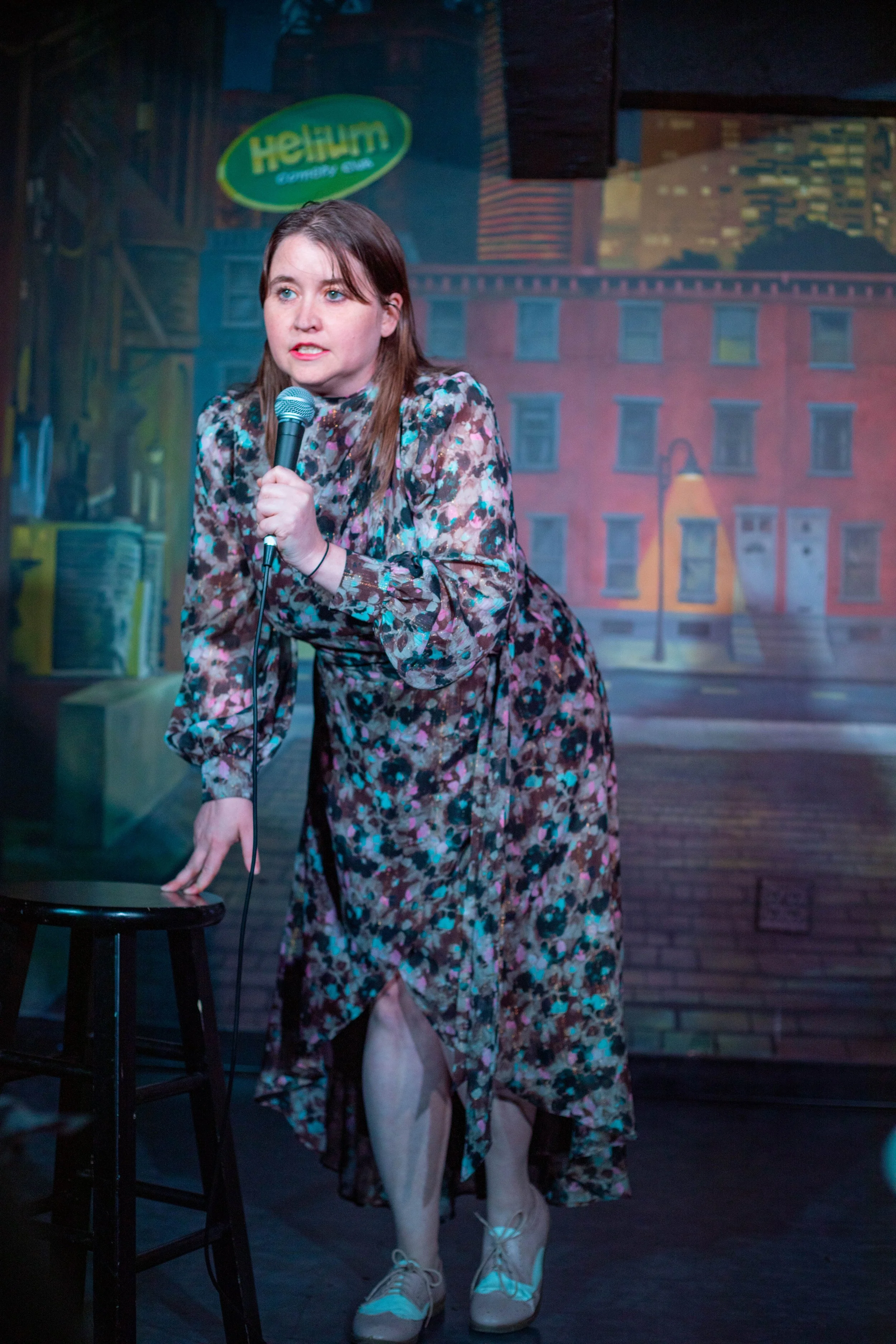 A woman on stage holding a microphone, leaning on a stool, in front of a painted backdrop depicting city buildings and a streetlamp.