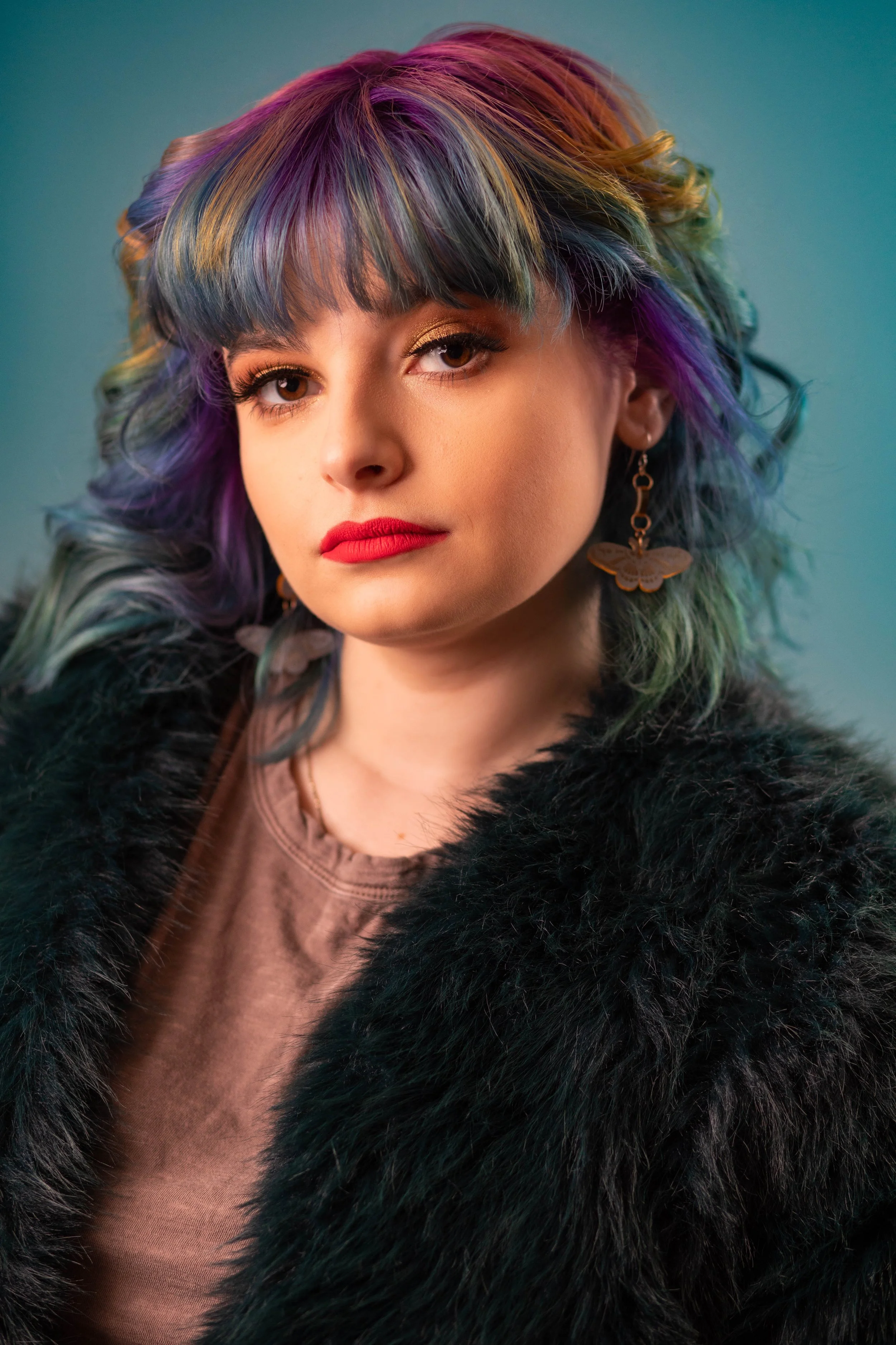 Portrait of a young woman with colorful, wavy hair in shades of purple, blue, green, and pink, wearing red lipstick, gold eye makeup, and butterfly earrings. She is dressed in a brown top and a black fur coat, with a neutral background.