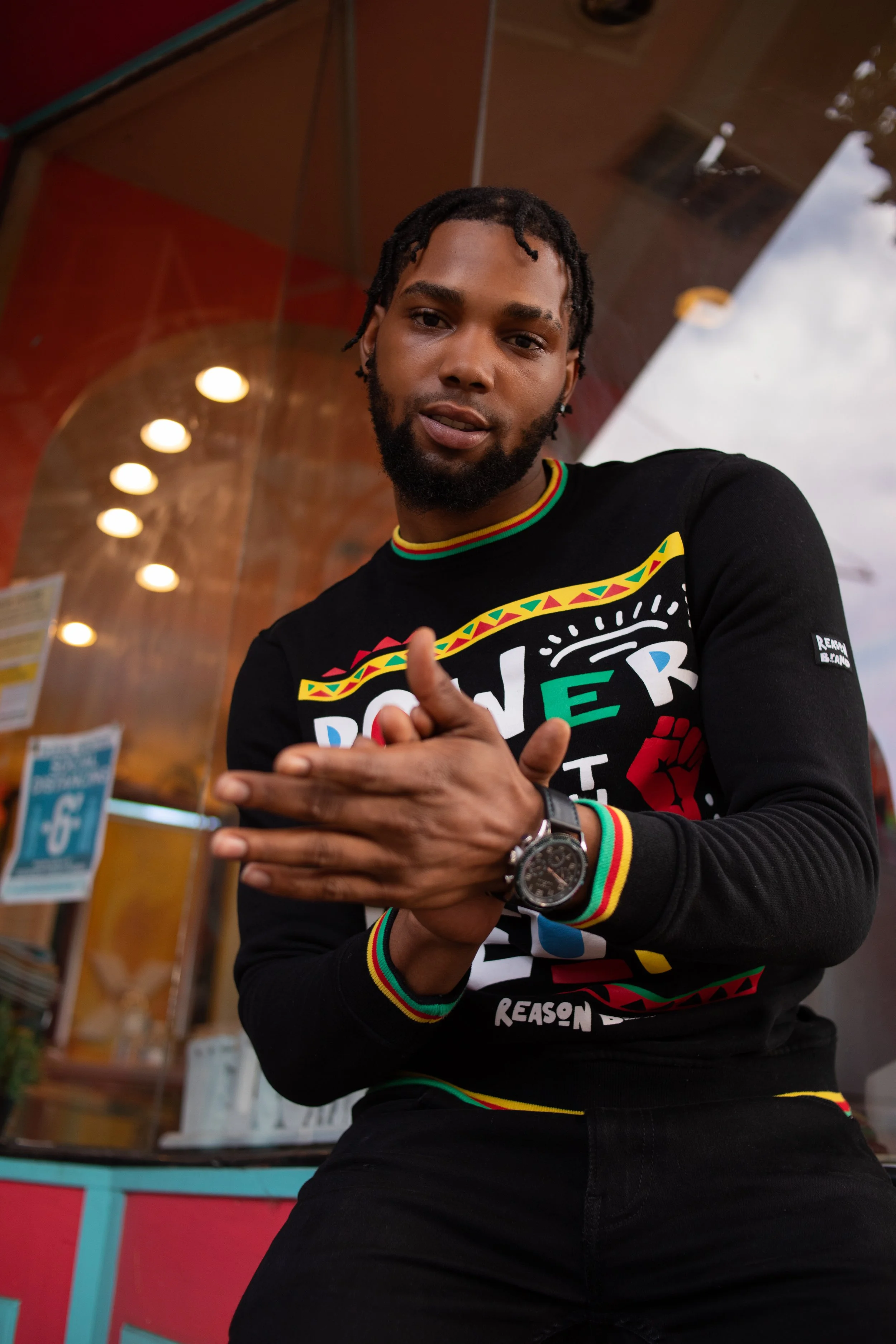 A young man with dark skin and dreadlocks, wearing a black shirt with colorful designs and text, is posing outdoors in front of a colorful building with round lights. He is making a gesture with his right hand and wearing a wristwatch.