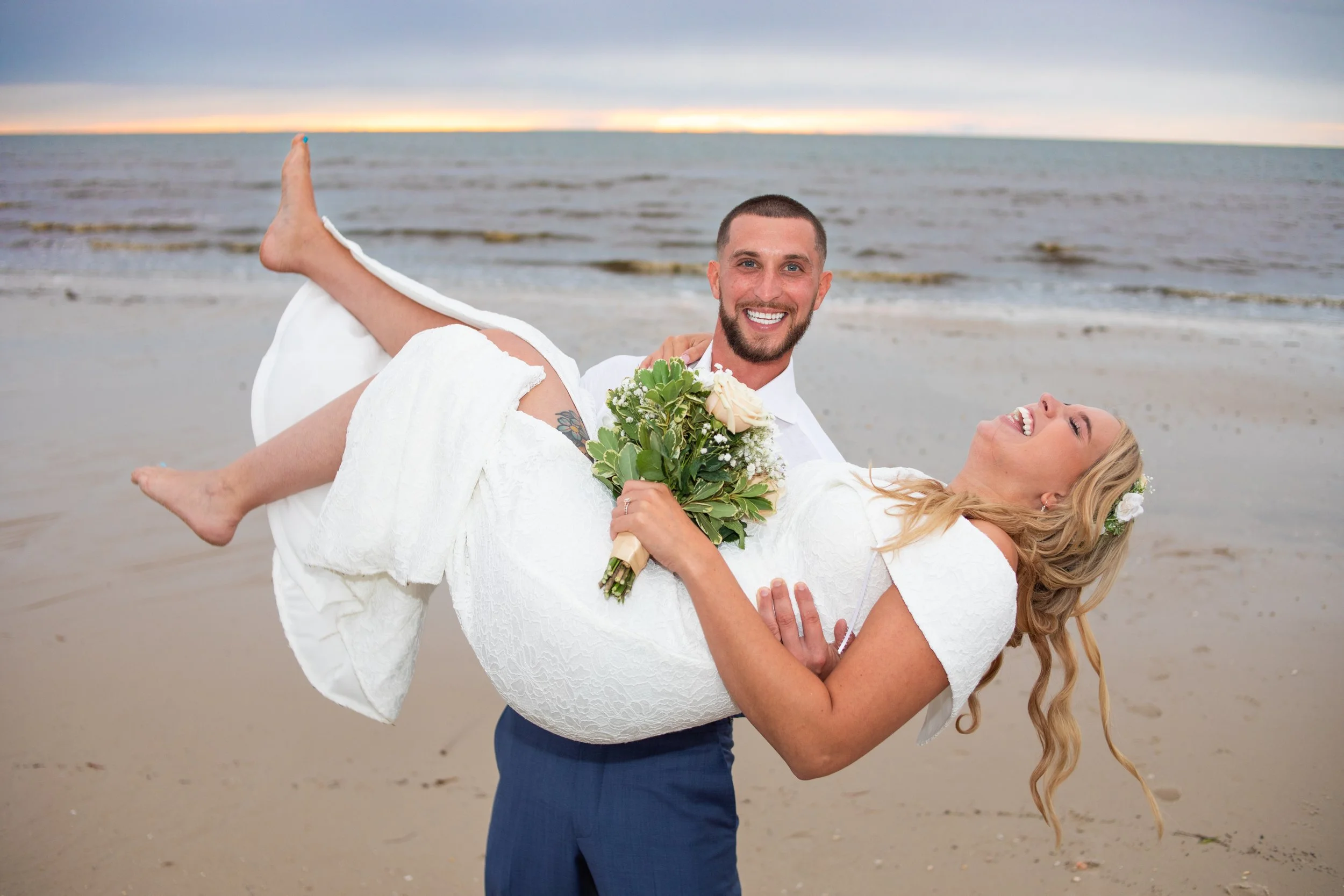 A newlywed couple at the beach, the groom in a white shirt and blue pants holding the bride, who is in a white dress and holding a bouquet of flowers, both smiling and laughing with the ocean and cloudy sky in the background.