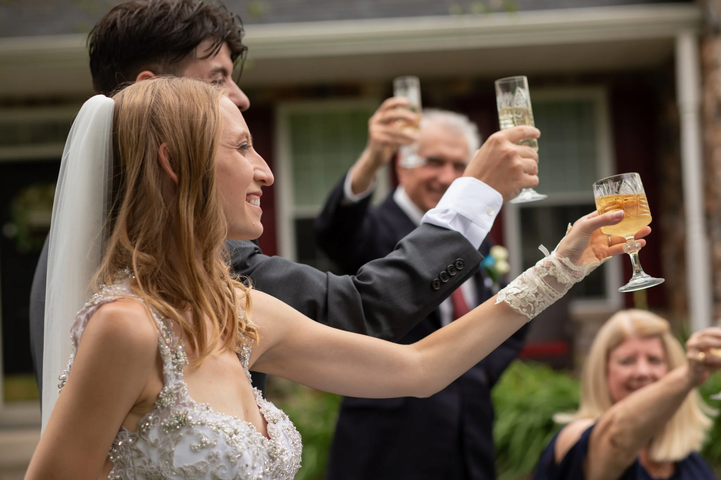 A bride and groom celebrating with family and friends at a wedding, raising glasses for a toast outdoors.