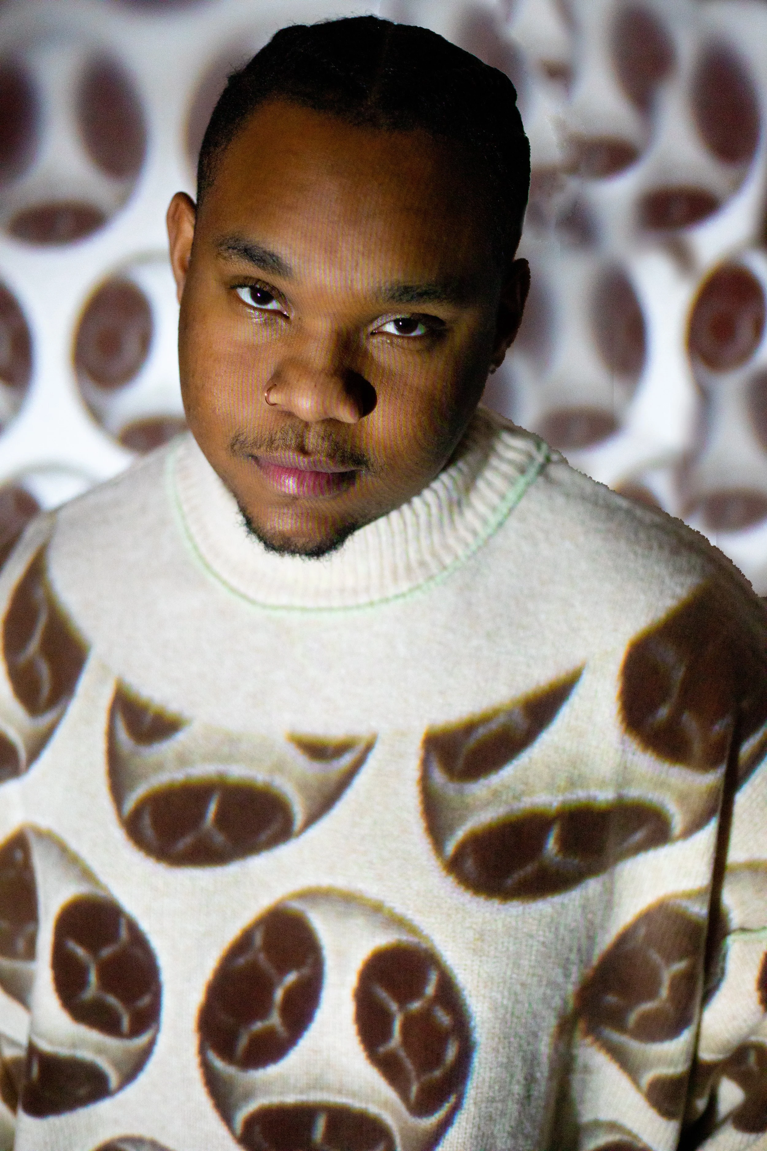 A young man with short hair and a goatee wearing a beige sweater with a tennis ball pattern, standing in front of a background with circular cutouts.