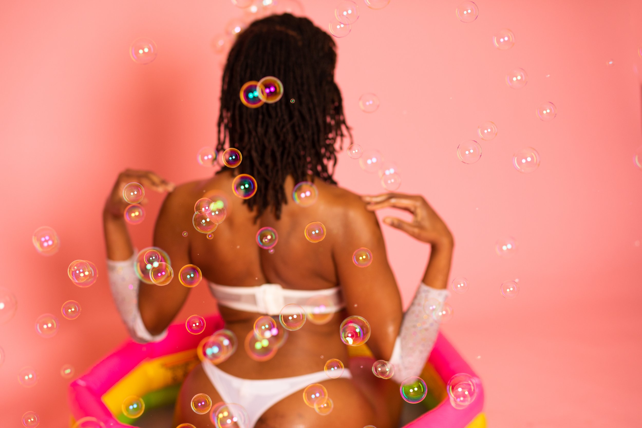 A woman with dreadlocks is sitting in a small pink pool with a yellow and green border, wearing white lingerie and long white lace gloves, surrounded by floating soap bubbles against a pink background.